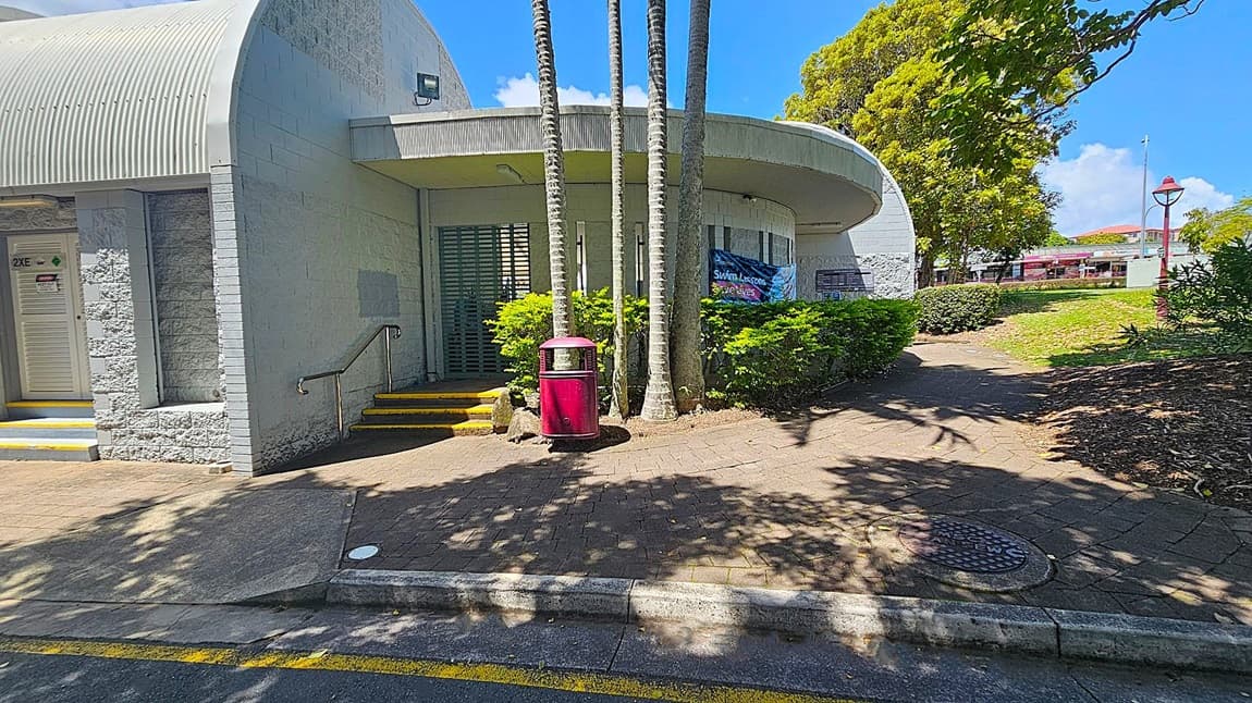 A building with a curved roof and light grey brick walls is on the left. A door marked "2XE" is visible with steps leading up to it. In the centre, several tall palm trees stand in front of a green metal gate, with a red bin nearby. Bushes and a banner advertising swim lessons are in front of the building. On the right, a paved path leads through a grassy area with trees and a red lamp post. Shadows of trees are cast on the ground.