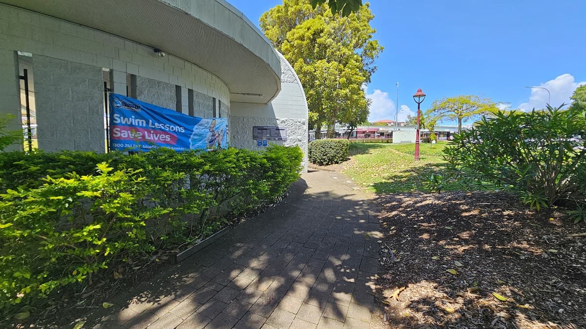 A curved building on the left with a banner reading "Swim Lessons Save Lives" is partially obscured by green shrubs. The path in the centre is paved with bricks, leading towards a grassy area on the right. A red lamp post stands on the grass, surrounded by trees and bushes. In the background, a few buildings are visible.