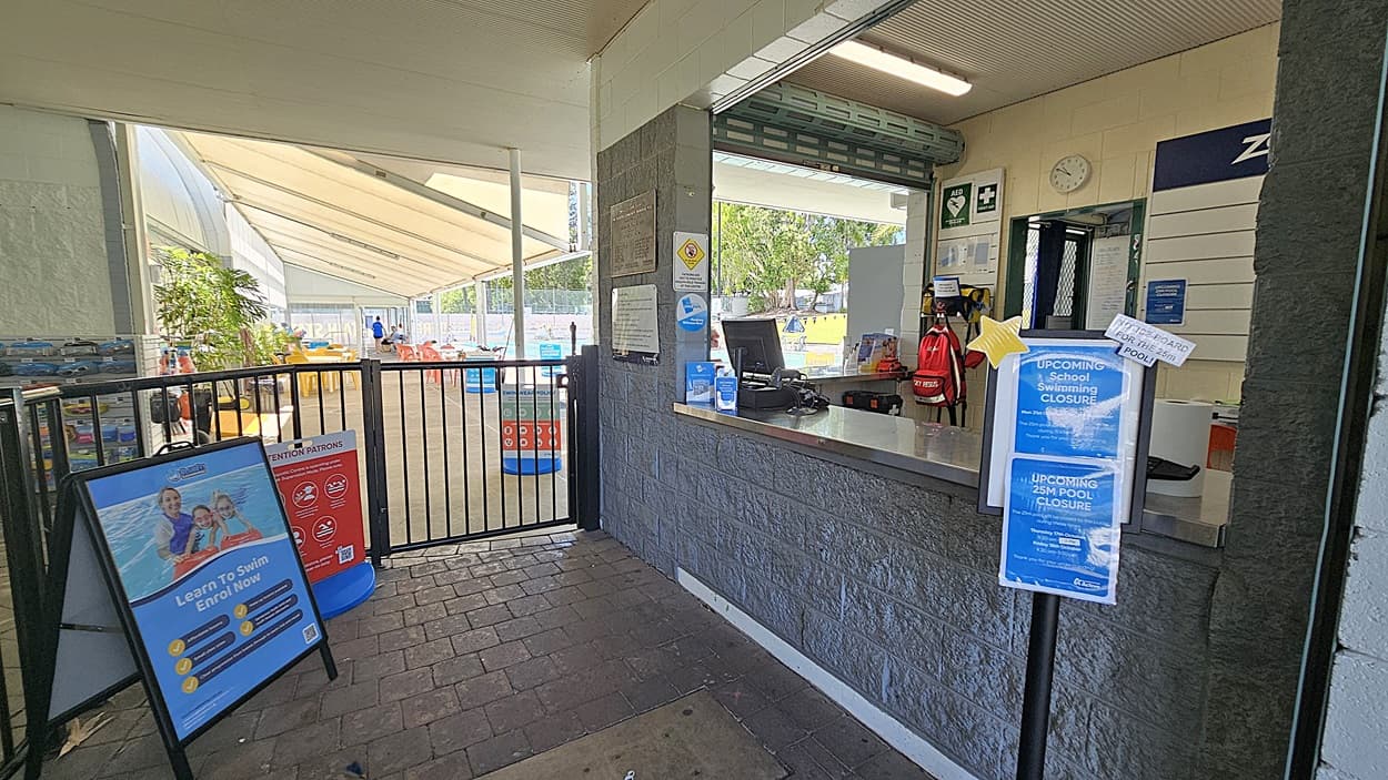 On the left, a gated entrance leads to a pool area with tables and chairs under a canopy. In the middle, a counter with a computer and various notices, including a sign about pool closures. On the right, a wall-mounted clock and first aid sign are visible. The floor is paved with dark bricks, and the ceiling is lined with light panels.