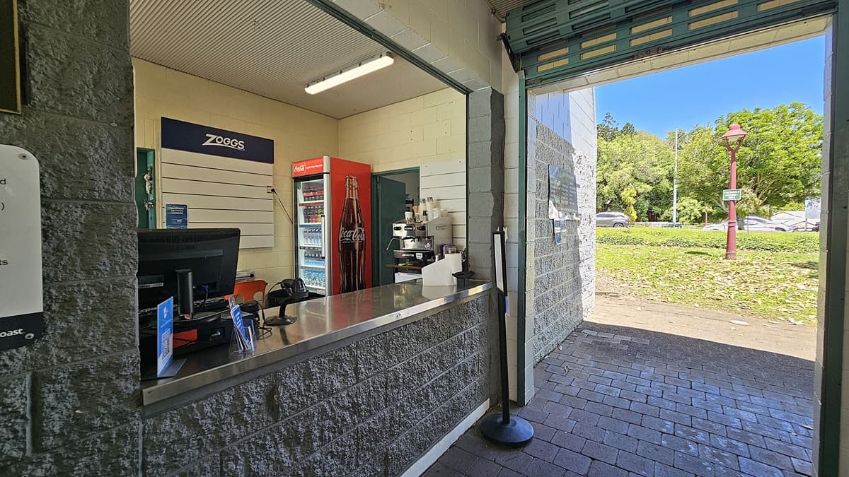 On the left, a counter with a computer and a Zoggs sign above. A red Coca-Cola fridge stands next to a coffee machine. In the middle, a metal counter extends towards the entrance. On the right, an open doorway leads outside to a grassy area with a red lamp post. The floor is paved with dark bricks, and the ceiling features a white panel with a fluorescent light.