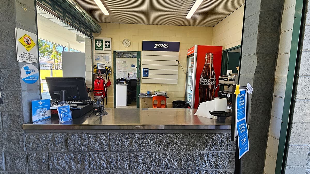 On the left, a computer and a sign about health measures are on a grey stone counter. In the middle, a wall with a Zoggs sign, a clock, and a first aid kit. On the right, a red Coca-Cola fridge and a coffee machine are visible. The floor is tiled, and the ceiling has white panels with fluorescent lights.