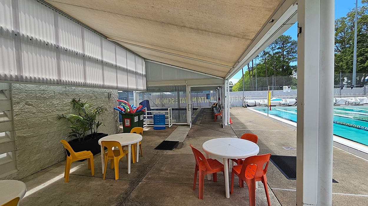 A covered outdoor area with a corrugated roof and a concrete floor. On the left, there are white plastic tables with yellow and orange chairs. A green wall with potted plants is visible. In the middle, a bin filled with colourful pool noodles is near a glass door. On the right, a swimming pool with lane dividers is visible, surrounded by a fence and trees in the background.