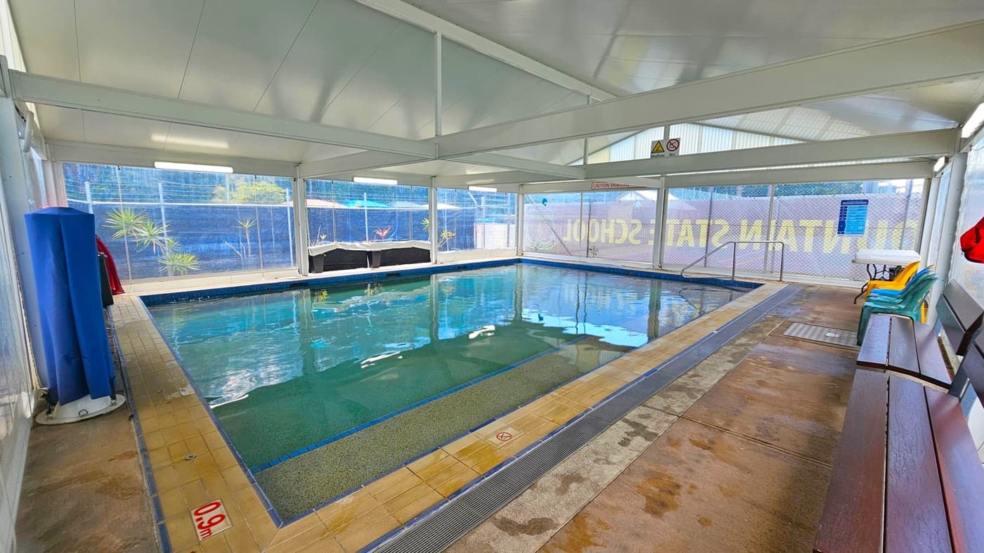 An indoor swimming pool with clear water, surrounded by a tiled deck. On the right, there are stacked plastic chairs and a wooden bench. The left side has a blue pool cover roller. Large windows with mesh fencing provide an outside view of plants and a blue fence. The ceiling is white with fluorescent lights, and the floor is wet with some puddles. Signs indicate pool depth and safety warnings.