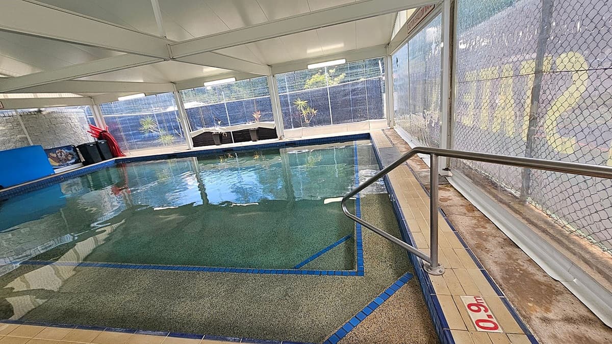 Indoor swimming pool with a tiled edge and a depth marker reading "0.9m" on the right. A metal handrail descends into the water. The pool is surrounded by large windows with a chain-link fence outside. On the left, there are red pool noodles and a black storage bin. The ceiling is white with fluorescent lights, and the floor is tiled. Some potted plants are visible outside the windows.