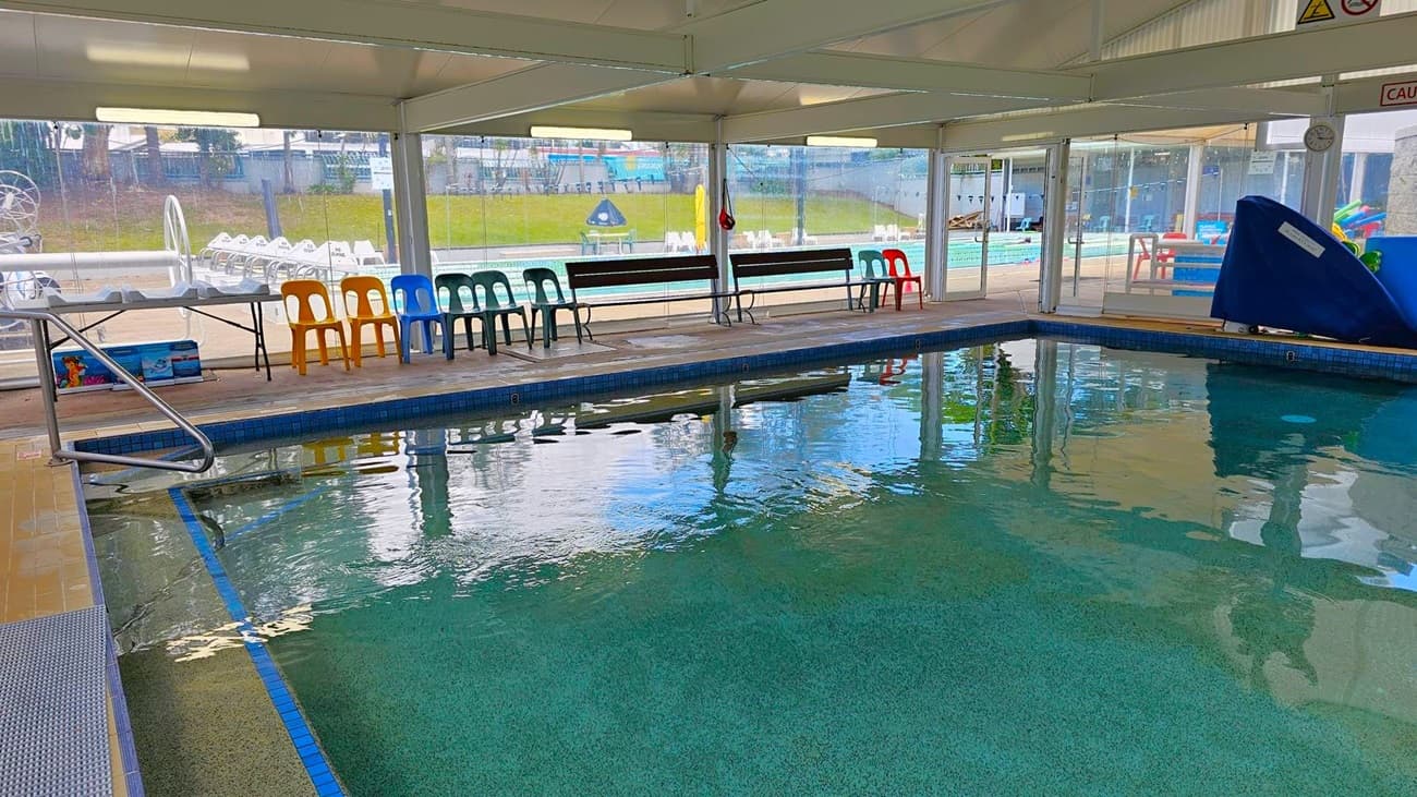 Indoor swimming pool with clear water, surrounded by a tiled deck. On the left, a metal handrail descends into the pool. Along the right side, there are colourful plastic chairs and a bench against a glass wall, providing a view of an outdoor area with more seating and a grassy hill. The ceiling is white with fluorescent lights, and a blue slide is visible near the pool's edge.