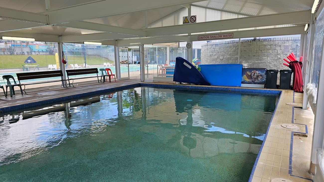 Indoor swimming pool with greenish water, surrounded by a tiled deck. On the left, there are black and red plastic chairs and benches. The right side has a blue slide and a stack of red pool noodles. Glass walls provide a view of an outdoor area with more seating and a grassy hill. The ceiling is white with fluorescent lights, and a sign reads "Caution Shallow Water."