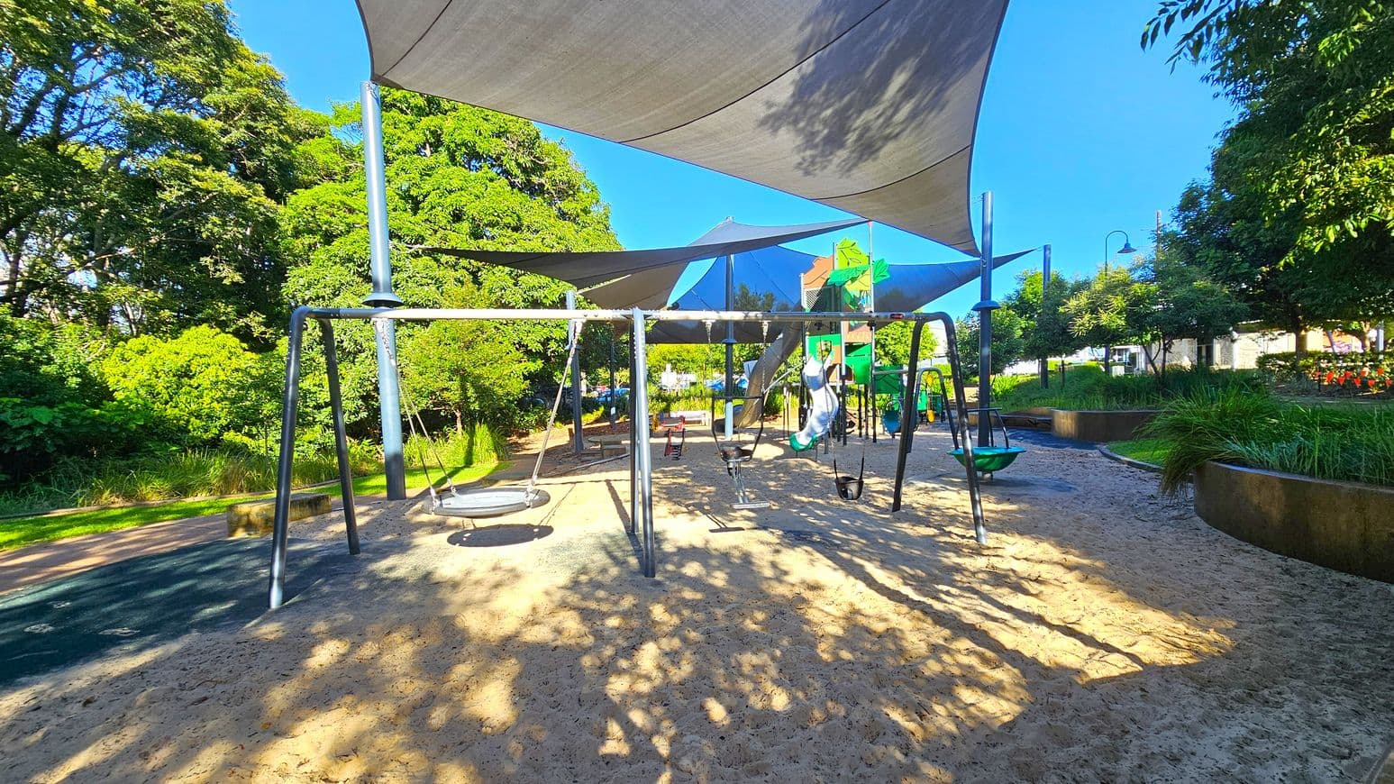 A playground with swings in the centre, set on a sandy surface. Overhead, there are large shade sails providing cover. To the left, lush green trees and bushes line the area. On the right, more greenery and a pathway are visible, along with some park benches. In the background, a play structure with slides and climbing elements can be seen. The area is well-lit, suggesting a sunny day.