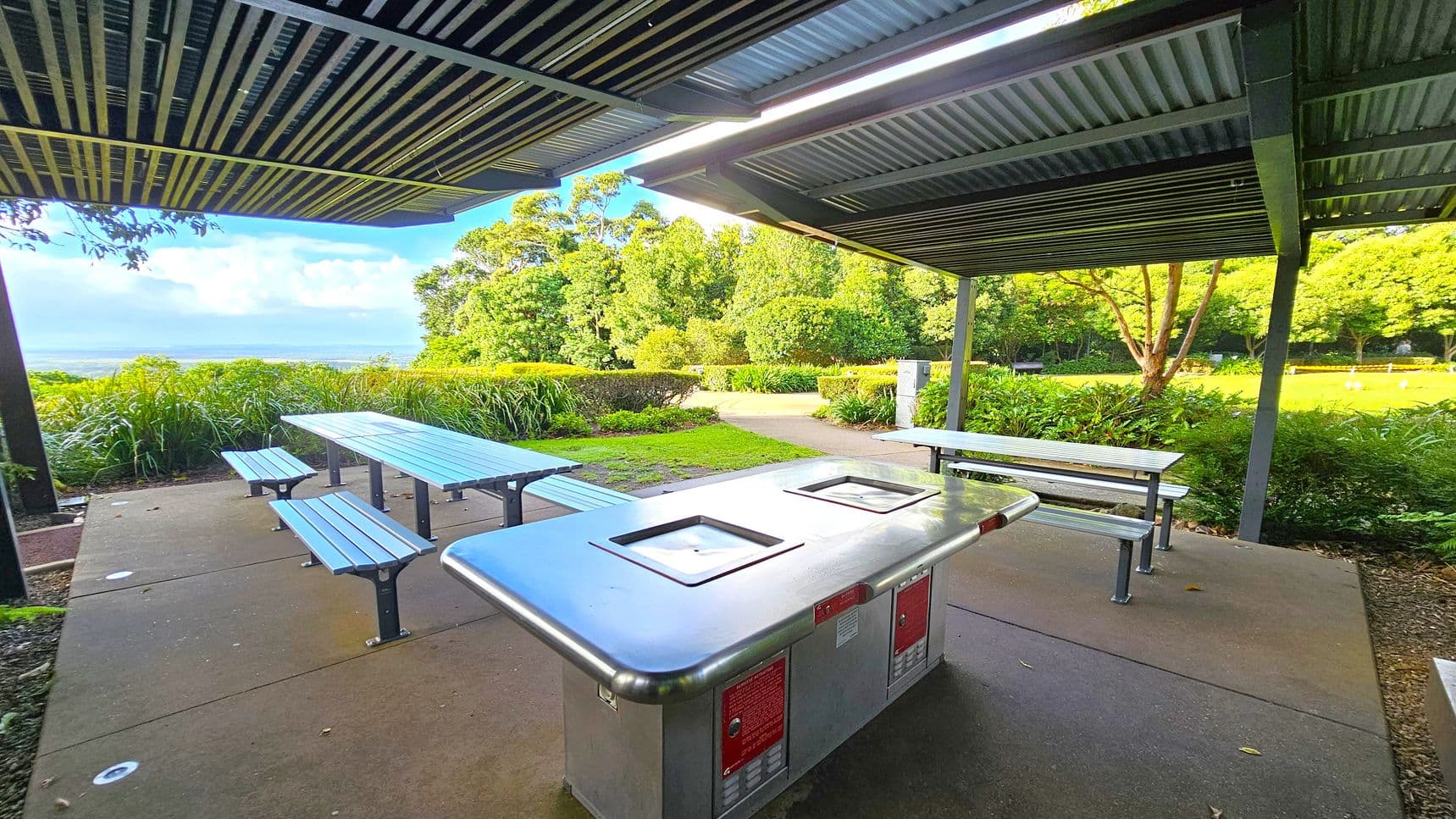 A covered outdoor area with a metal barbecue grill in the foreground. To the left, there are picnic tables with benches. The ground is paved with concrete, and the ceiling is made of corrugated metal. In the background, lush greenery and trees are visible, with a pathway leading through the grass. The area is surrounded by bushes and plants, creating a serene park setting.