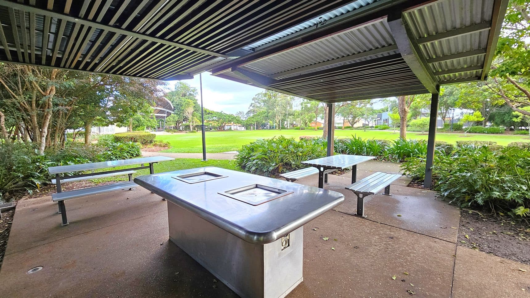 A covered outdoor area with a metal roof and a concrete floor. In the foreground, there is a stainless steel barbecue grill. To the left and right, there are metal picnic tables with benches. The background features a grassy park with trees and shrubs, and a path leading through the greenery.
