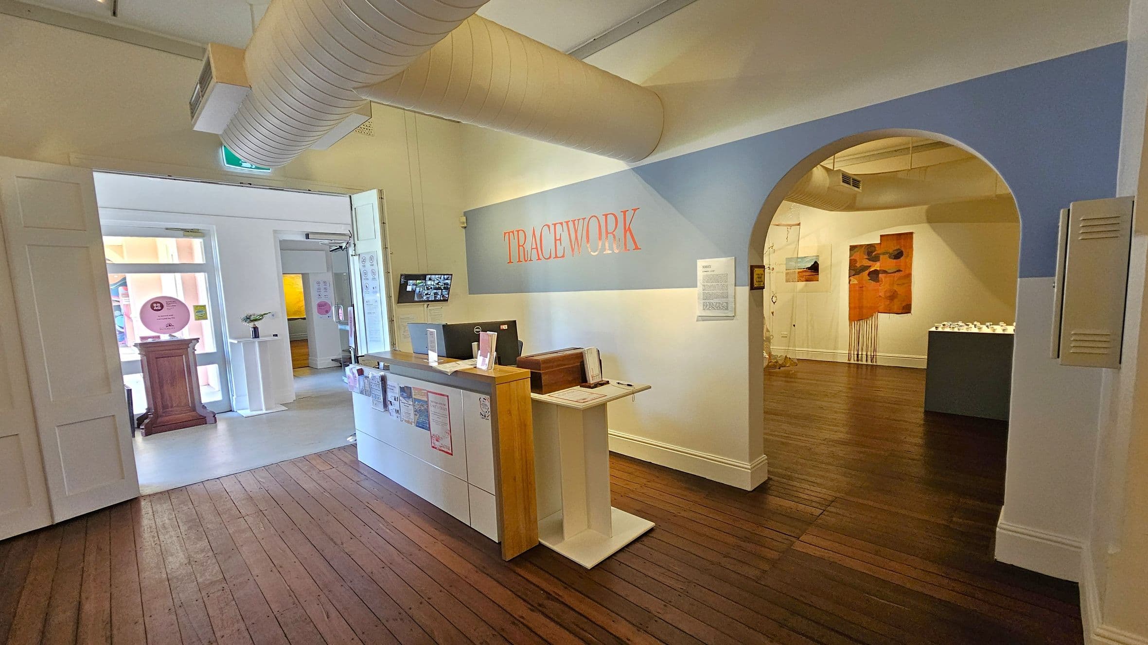 A gallery interior with wooden flooring. On the left, there's a reception desk with brochures and a computer. Behind it, a doorway leads to another room with a wooden pedestal and a circular sign. Above, a large duct runs across the ceiling. On the right, an arched entrance displays artwork on the walls, including a large orange piece. The word "TRACEWORK" is prominently displayed on the wall above the archway.