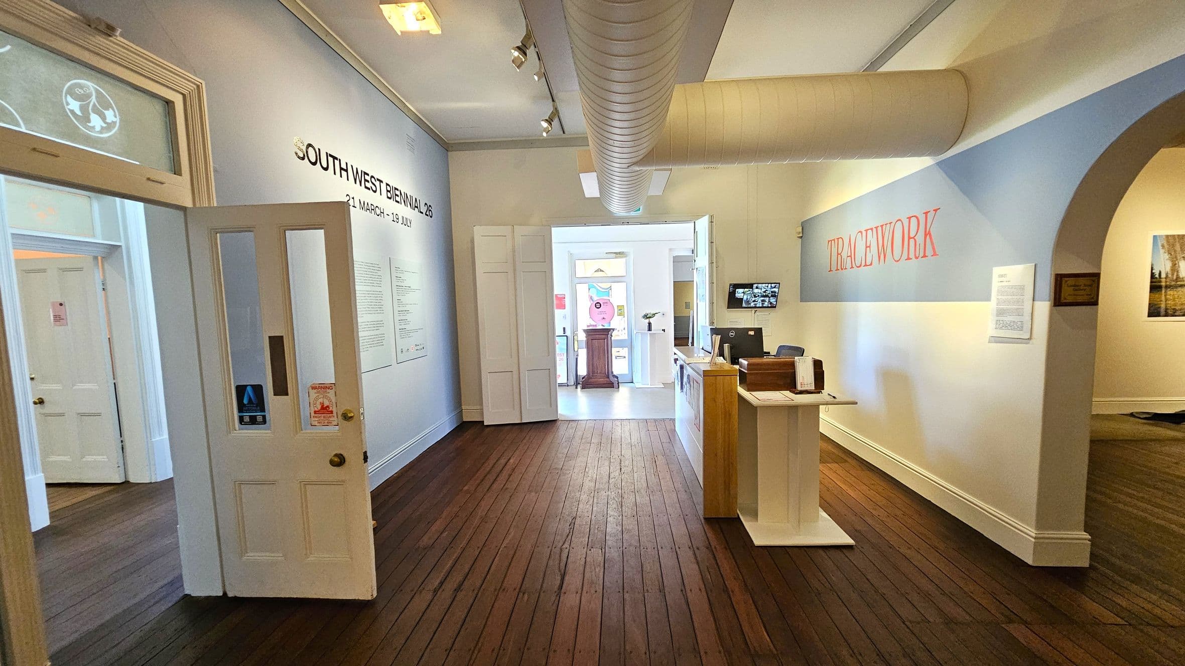 A gallery interior with wooden flooring and light-coloured walls. On the left, a door is open, revealing another room. The wall displays text about the "South West Biennial 26" exhibition. In the centre, a doorway leads to a bright area with a wooden podium. On the right, a reception desk is positioned under a wall with the word "TRACEWORK" in red. An archway leads to another room with artwork visible. A large duct runs along the ceiling.