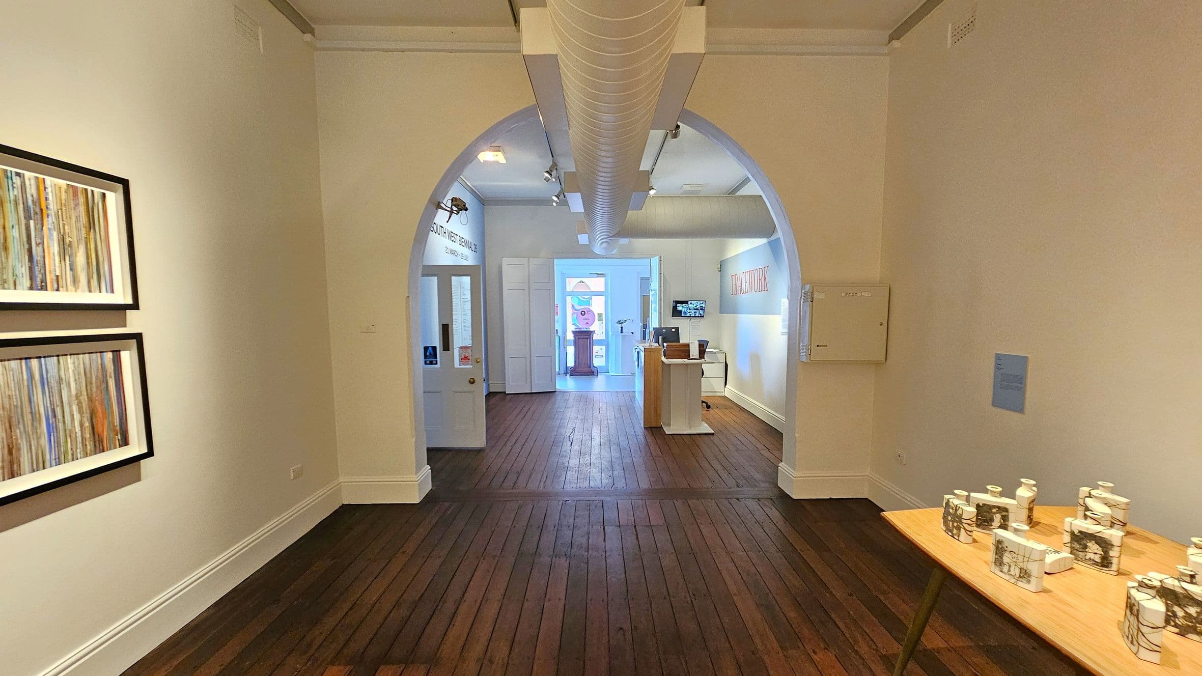 A gallery interior with wooden flooring and white walls. On the left, two framed abstract artworks hang on the wall. In the centre, an arched doorway leads to another room with a wooden pedestal and a colourful artwork visible in the distance. On the right, a wooden table displays several ceramic pieces with intricate designs. A ventilation duct runs along the ceiling.