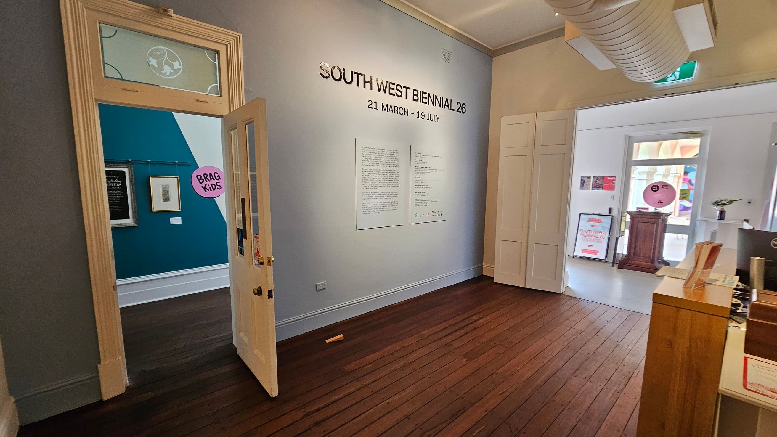 A room with wooden flooring and light-coloured walls. On the right, a wall displays "SOUTH WEST BIENNIAL 26" with dates and text below. A doorway on the left leads to another room with teal walls, framed artwork, and a "BRAG KIDS" sign. In the background, a small wooden podium and a sign are visible near a window. A desk with brochures is in the foreground, and a large air duct is on the ceiling.