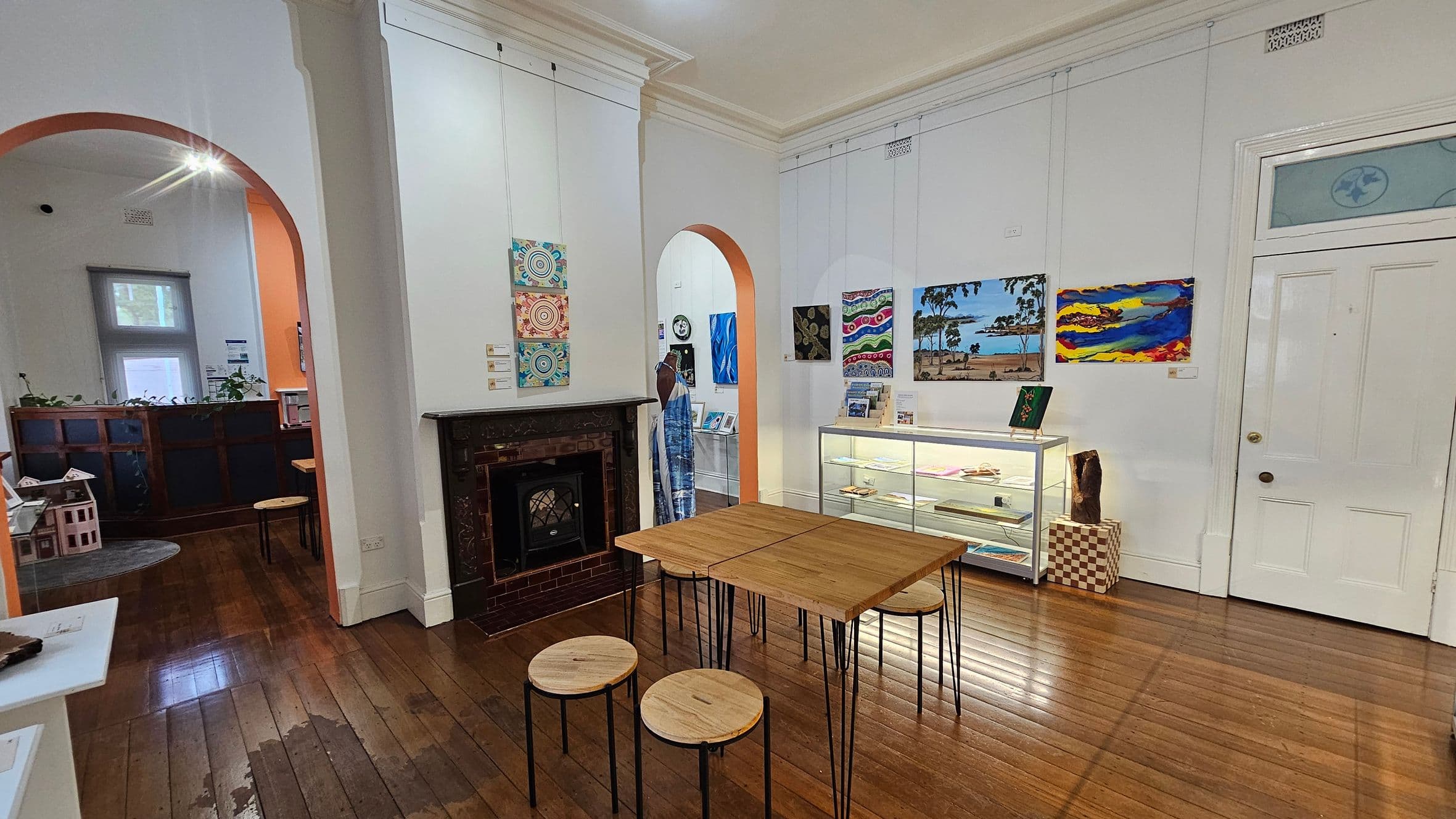 A room with wooden flooring and a high ceiling. In the centre, there is a wooden table with round stools. On the right, a glass display case holds various items, and colourful paintings hang on the wall above it. A white door is visible next to the display. On the left, a fireplace with a dark mantel is set into the wall, and an archway leads to another room with a window and plants on a cabinet.