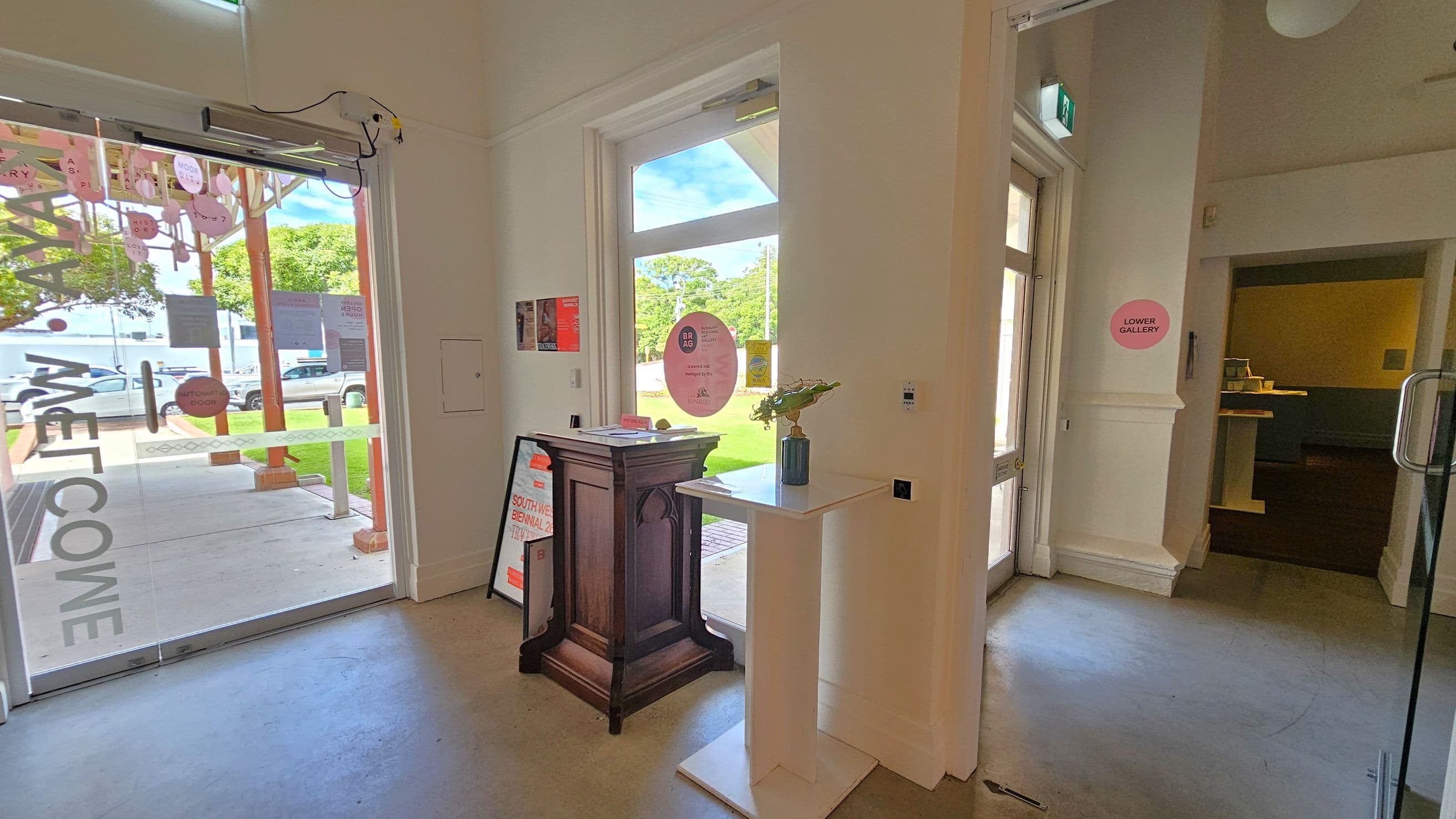 A bright interior space with a glass entrance door on the left, displaying the word "WELCOME" and various circular stickers. Outside, a green lawn and trees are visible. Inside, a wooden stand with informational signs is near the door. A white pedestal with a vase of flowers is positioned in the centre. On the right, a doorway leads to a room marked "LOWER GALLERY". The floor is smooth and grey, and the ceiling is white with a visible light fixture.