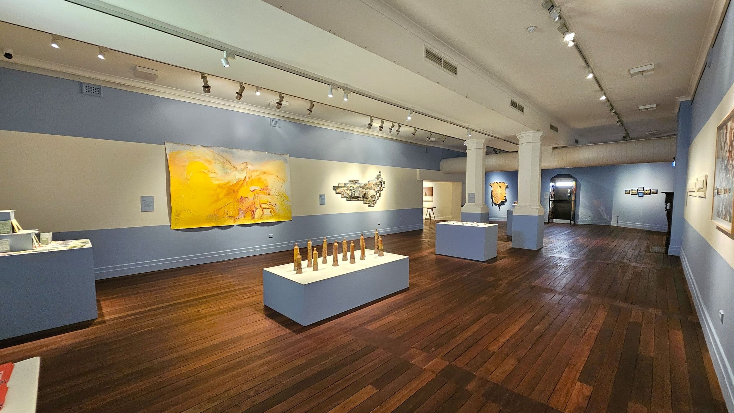 An art gallery with wooden flooring and a high ceiling with track lighting. On the left, a large yellow painting is displayed on a blue and cream wall. In the centre, a white pedestal holds several wooden sculptures. Further back, another pedestal displays more artwork. The right wall features additional paintings. A white column stands near the centre, and an archway in the background leads to another room.