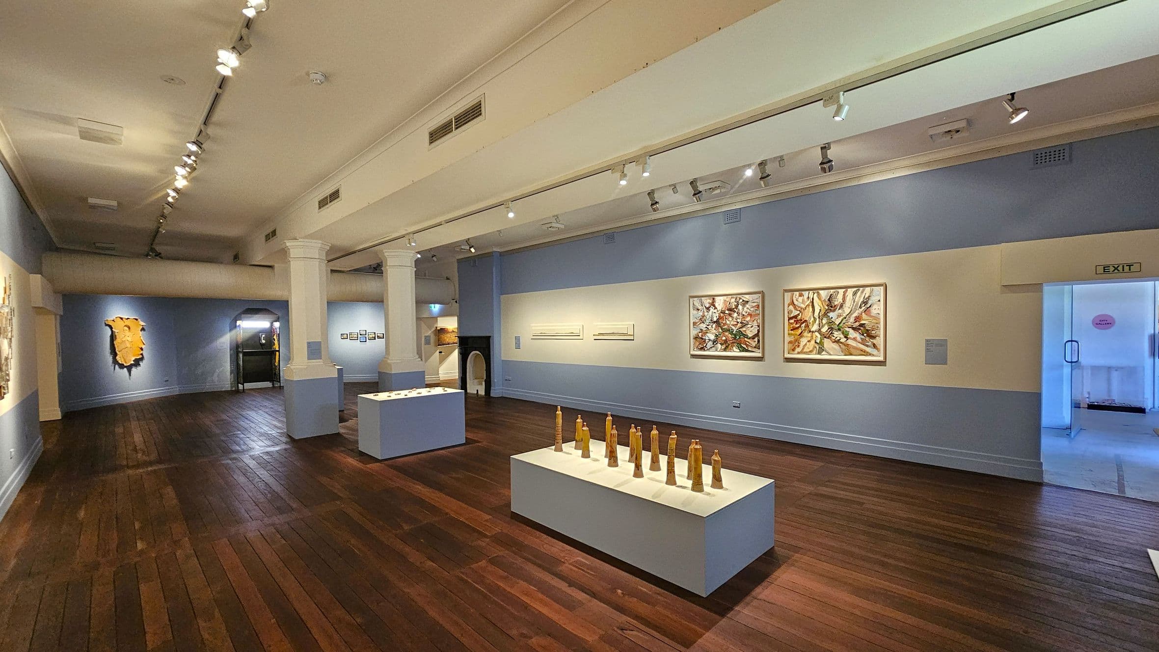 An art gallery with wooden flooring and a white ceiling featuring track lighting. On the right, there are two large abstract paintings on a blue wall, with an exit door nearby. In the centre, a display of wooden sculptures is arranged on a white pedestal. On the left, a textured artwork is mounted on the wall. Two large white columns divide the space, and another pedestal with small items is visible further back.