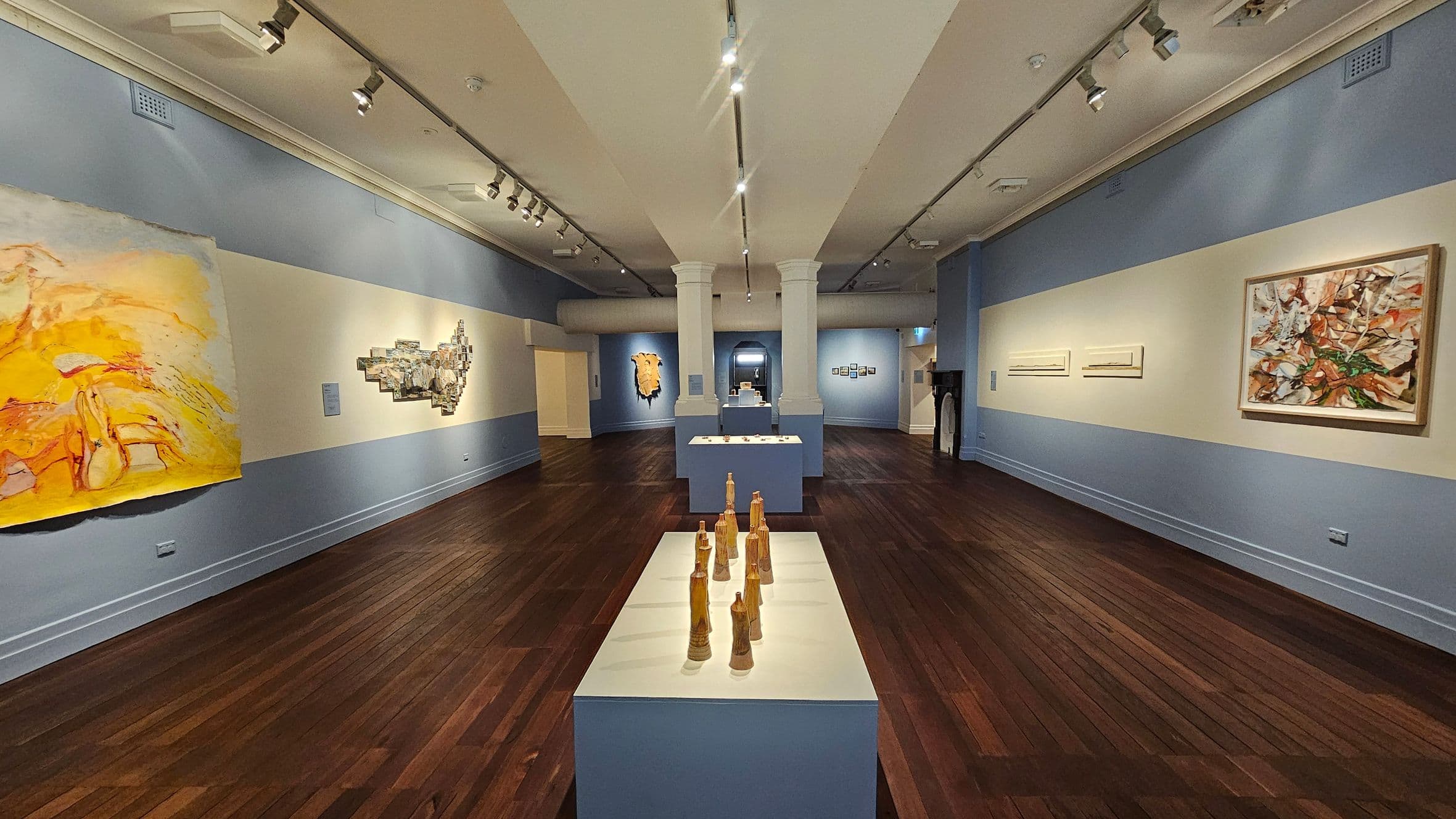 An art gallery with wooden flooring and a high ceiling featuring track lighting. In the centre, a display table holds several wooden sculptures. The left wall displays a large abstract painting with yellow and orange hues, and a textured artwork. The right wall has a framed abstract painting with earthy tones and two smaller rectangular pieces. The background shows more artworks and a doorway leading to another room.