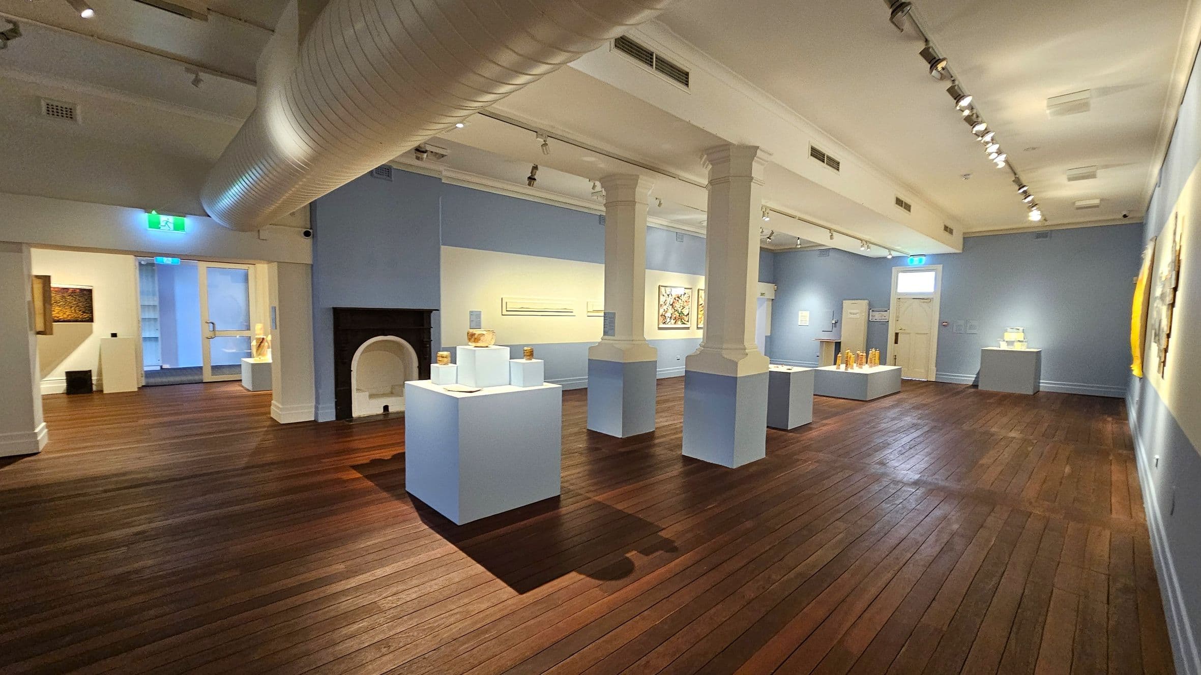 A spacious gallery with wooden flooring and light blue walls. In the centre, there are white display pedestals with various artworks. Two large white columns support the ceiling, which features a prominent air duct. On the left, a black fireplace is visible, and an exit sign is above a doorway. The right wall displays framed artworks. The room is well-lit with track lighting, and there are additional artworks on pedestals towards the back.