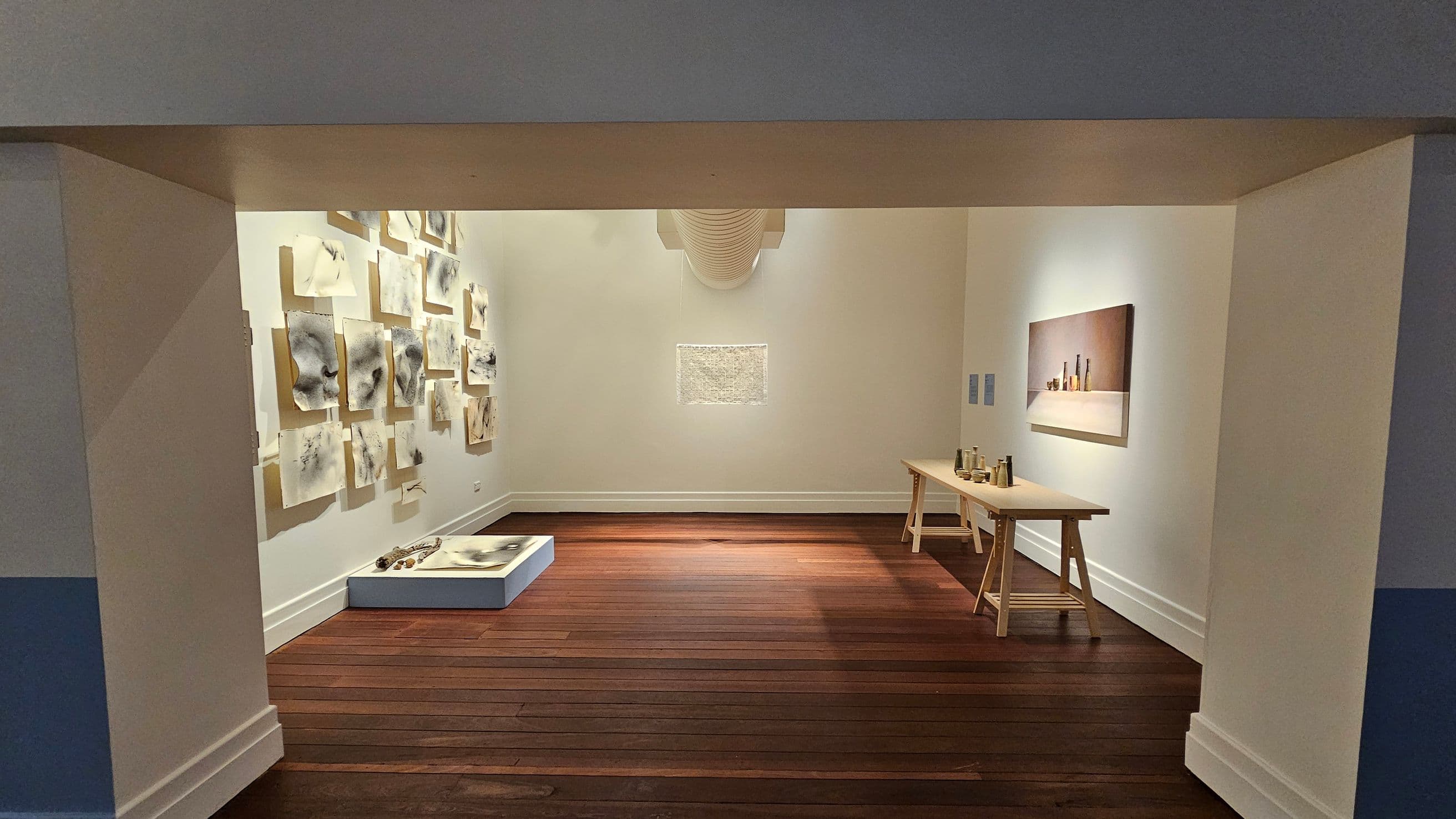 A gallery room with wooden flooring. On the left wall, numerous abstract artworks are displayed, arranged in a grid pattern. Below them, a low platform holds additional items. The right wall features a long wooden table with various objects on top and a painting of bottles hanging above. The back wall has a single framed piece, with a duct visible above it. The ceiling is plain and light-coloured.