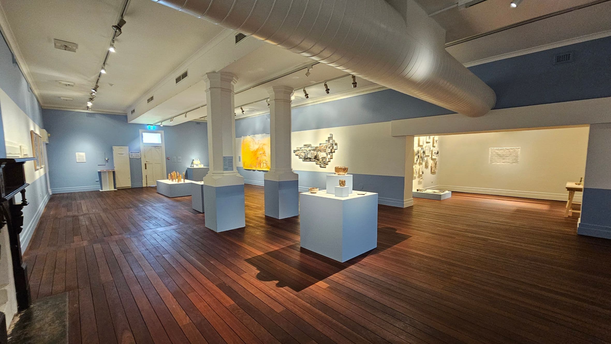 A spacious gallery with wooden flooring and light blue walls. In the centre, there are white display pedestals showcasing various artworks. Two large columns support the ceiling, which features exposed ductwork and track lighting. On the left, framed artworks are mounted on the wall. The right side opens into another room with additional displays. A small window and a door are visible at the back.