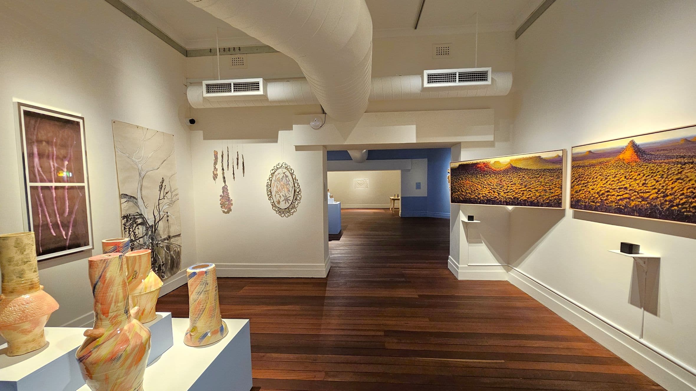 An art gallery with wooden flooring and a white ceiling featuring exposed ductwork. On the left, there are ceramic vases on a pedestal and framed artworks on the wall. In the centre, the wall displays hanging art pieces. On the right, two large landscape paintings are mounted. The background shows another room with additional artworks and a blue accent wall.