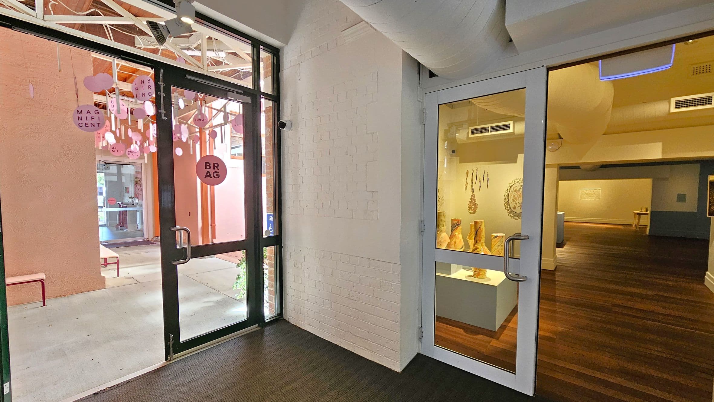 On the left, a glass door with a black frame opens to an outdoor area with pink walls and hanging decorations featuring words like "MAGNIFICENT" and "INSPIRING". A bench is visible outside. On the right, another glass door leads to an indoor gallery with wooden flooring, displaying sculptures on pedestals and wall art. The ceiling has exposed ducts and lighting fixtures. The floor inside is carpeted near the entrance.