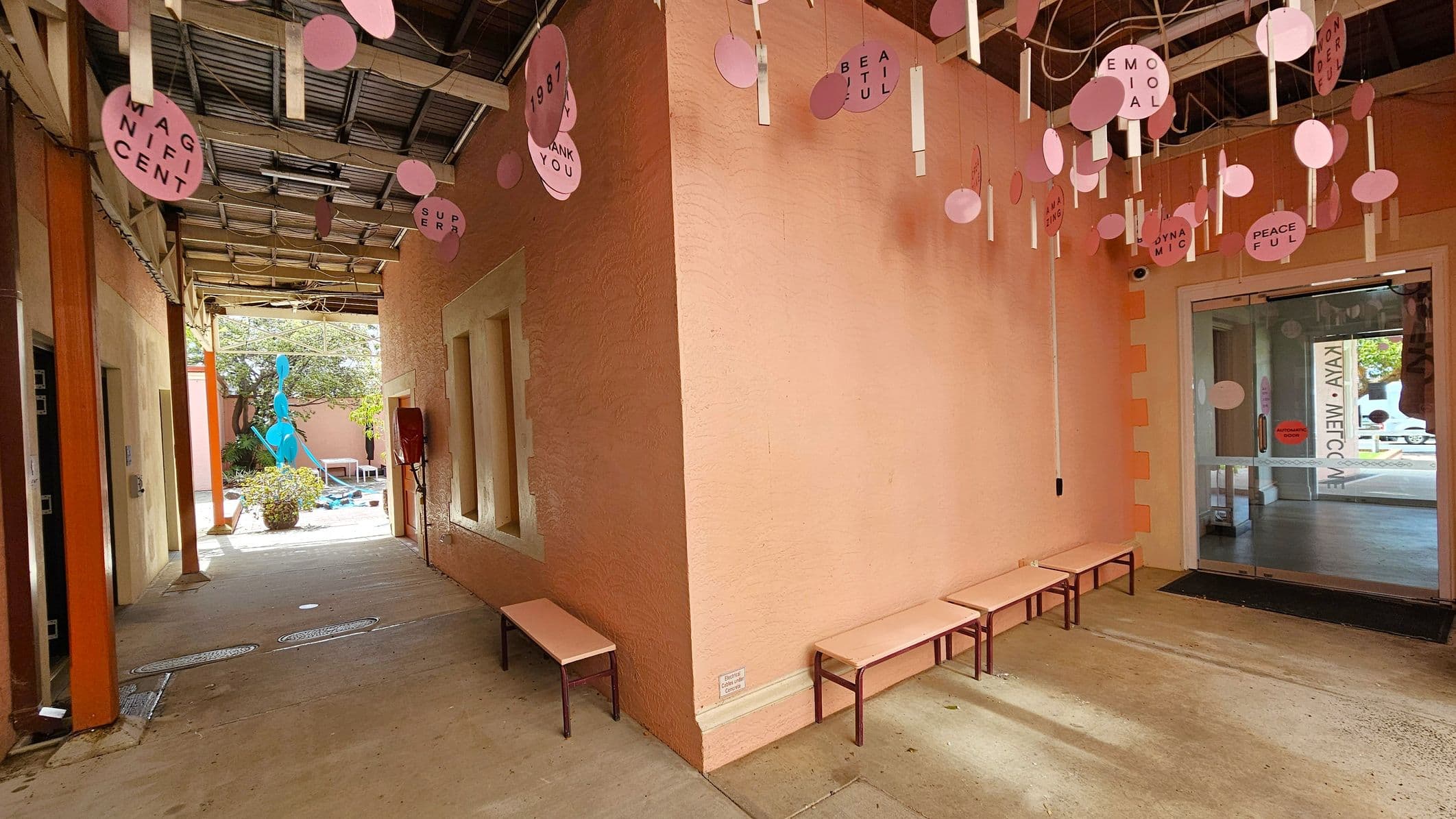 A corridor with peach-coloured walls and a concrete floor. On the right, there are several benches against the wall. Hanging from the ceiling are pink circular decorations with words like "MAGNIFICENT" and "BEAUTIFUL". On the left, the corridor leads to an outdoor area with a blue abstract sculpture and some greenery. The ceiling is open with visible beams. At the far end, there is a glass door with text on it.