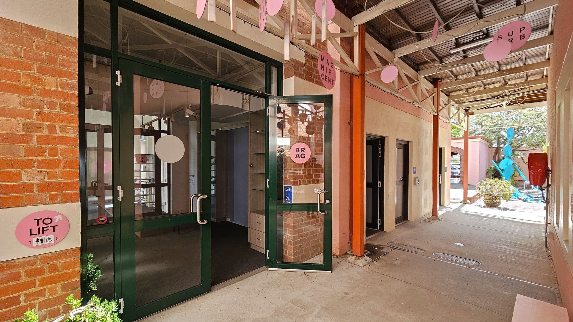 A corridor with a brick wall on the left featuring a glass door with metal handles, partially open. A pink sign on the wall indicates "TO LIFT" with symbols for accessibility. Above, pink circular decorations hang from the ceiling. On the right, a concrete path leads to an outdoor area with a blue abstract sculpture and greenery. The ceiling is metal with exposed beams, and a red fire hose reel is mounted on the wall.