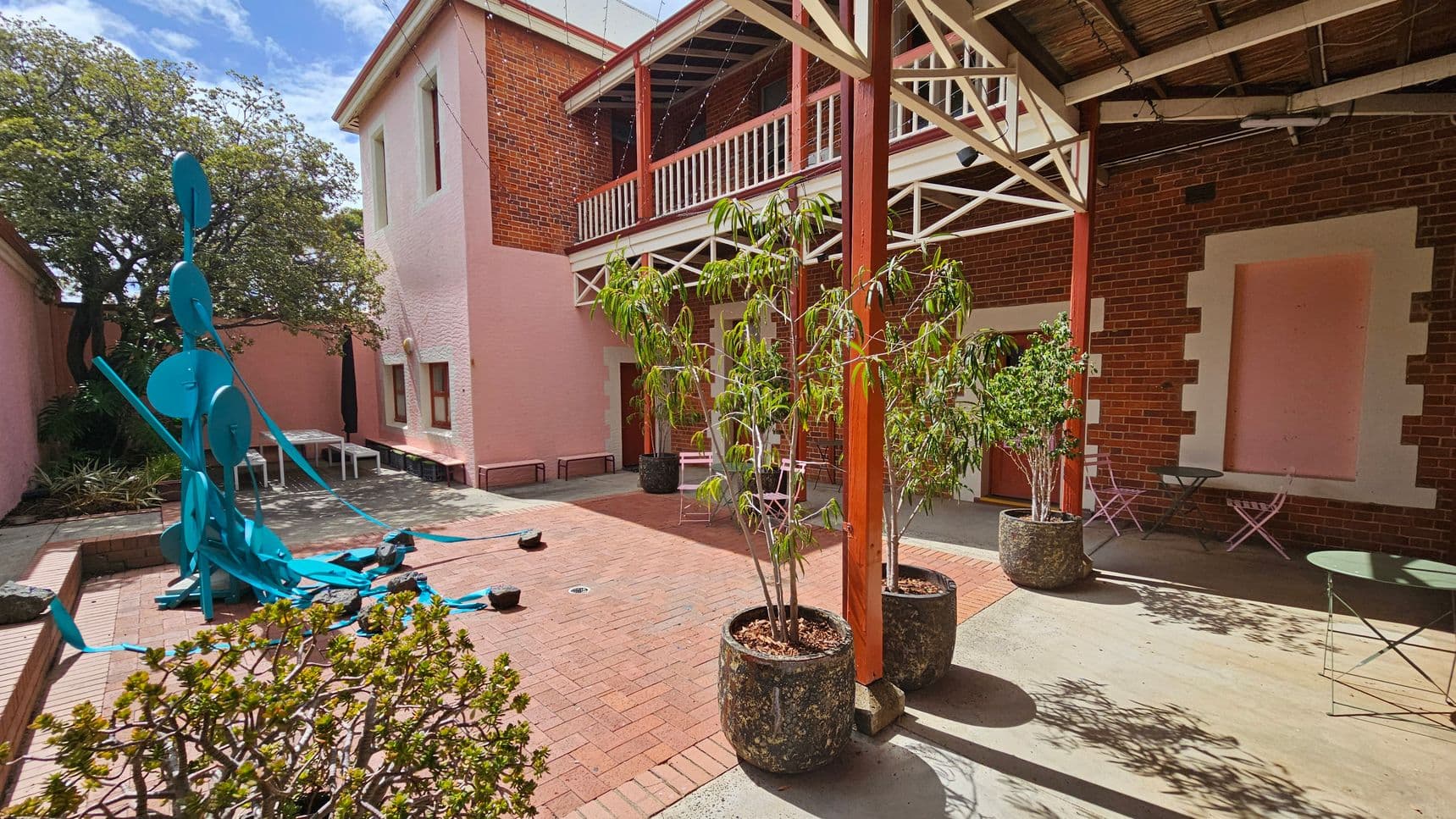 A courtyard with a brick floor and a mix of pink and red brick walls. On the left, a large blue abstract sculpture stands near a tree. In the centre, potted plants are placed on the ground. On the right, there are small tables and chairs against the wall. The building has a balcony with white railings and a wooden roof structure above.