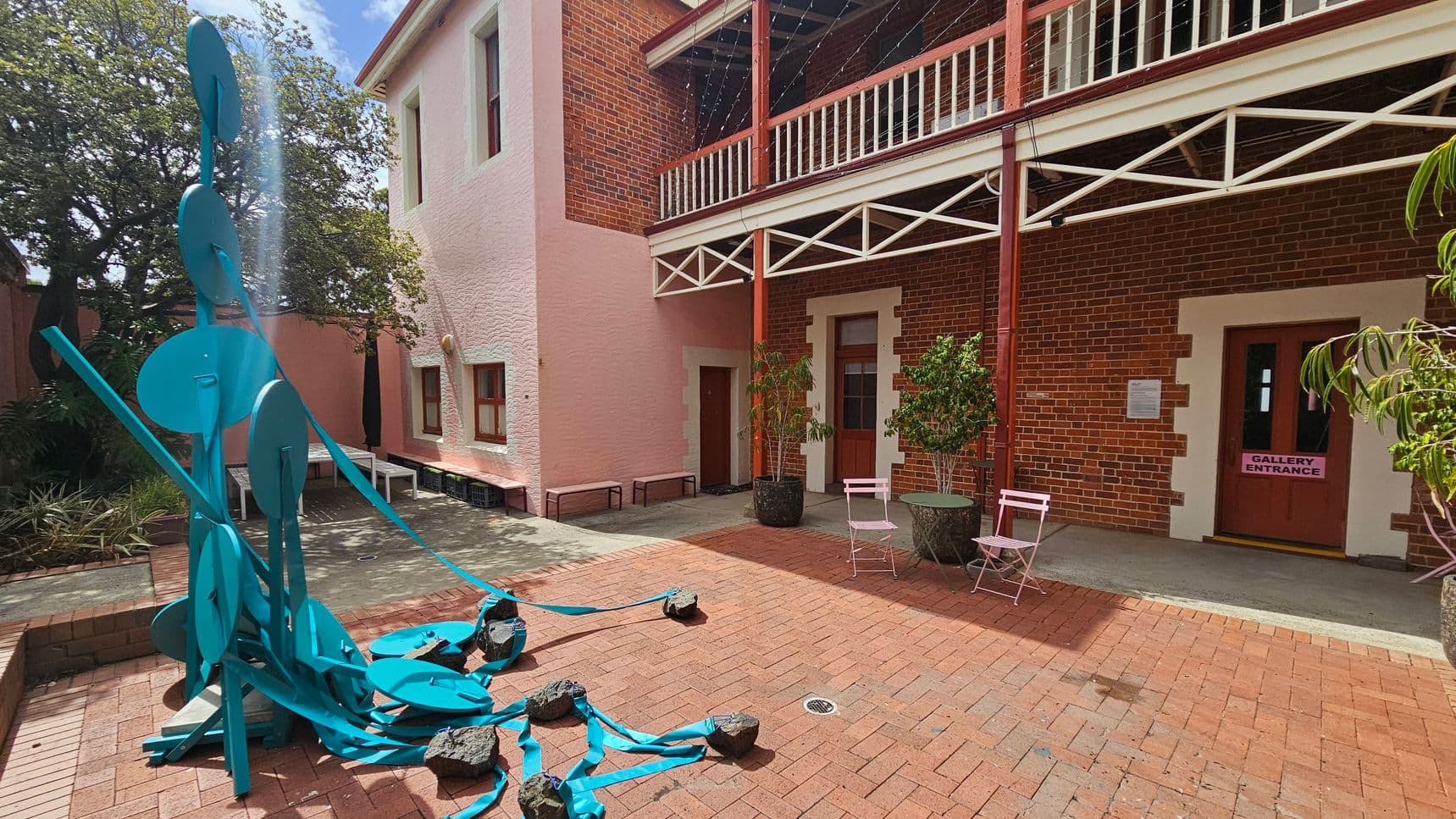 A courtyard with a brick floor and a two-storey building. On the left, a blue abstract sculpture with circular elements and ribbons is surrounded by rocks. The building has a pink and red brick facade with a balcony supported by white beams. Two potted plants and a small seating area with pink chairs and a table are near the entrance. The door on the right has a sign reading "Gallery Entrance." Trees and benches are visible in the background.