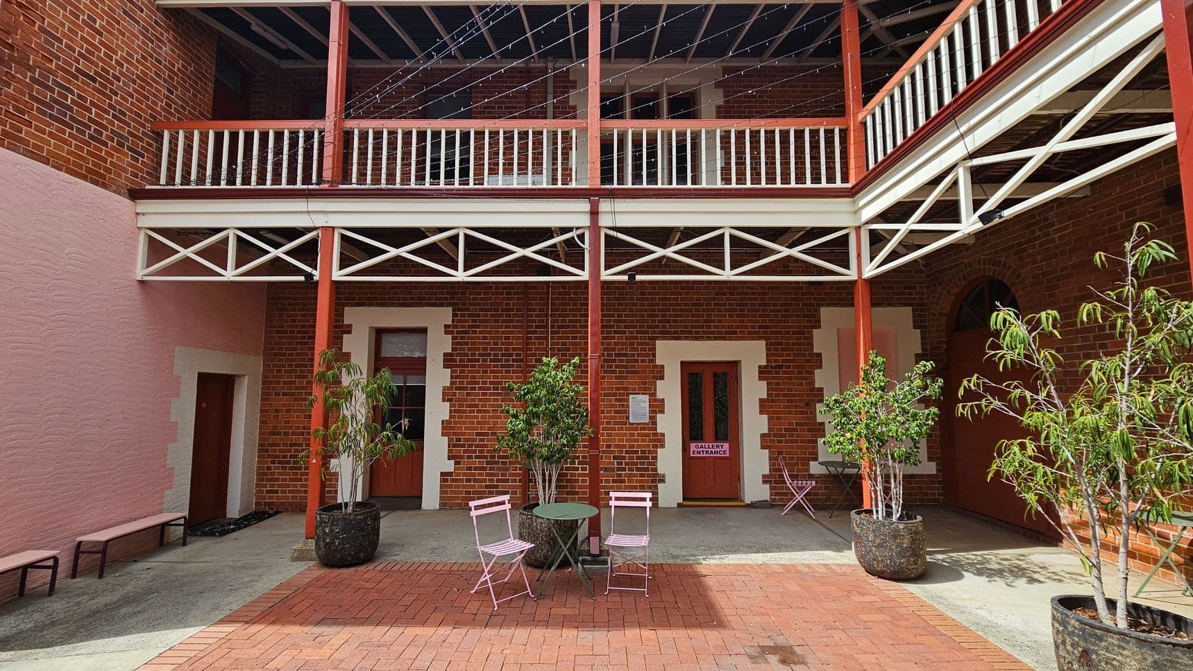 A courtyard with red brick walls and a two-level wooden balcony featuring white railings and supports. The ground is paved with red bricks. In the centre, there are two pink metal chairs and a small round table. Potted plants are placed around the area, including two large pots with leafy trees. A sign on one door reads "Gallery Entrance." The ceiling of the balcony is open with visible beams.