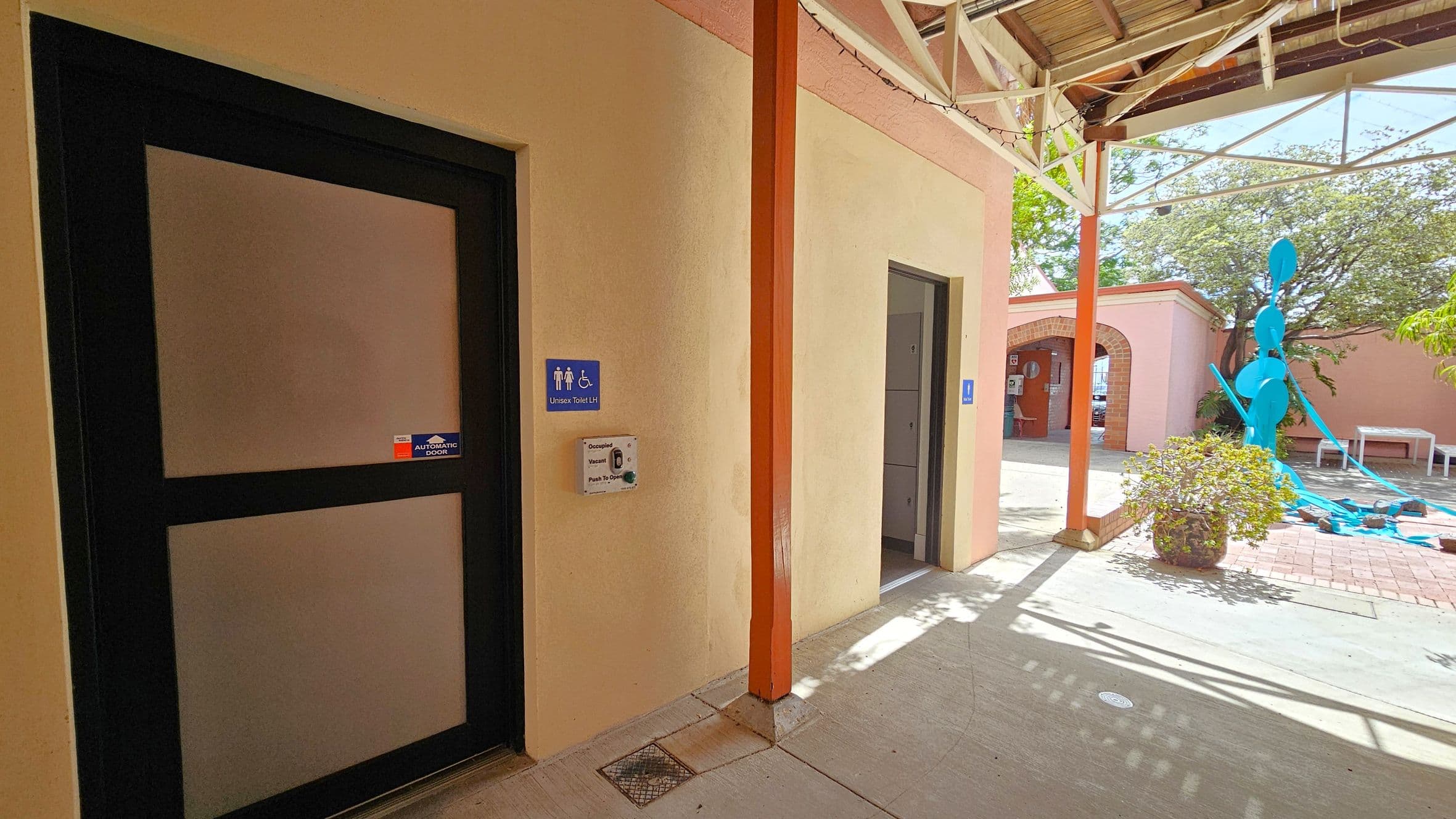 A corridor with a beige wall on the left featuring a black-framed door marked as a unisex toilet with accessibility signs. An automatic door button is mounted beside it. Further along, another door is visible. On the right, an open area with a large blue sculpture and a potted plant. The ground is a mix of concrete and brick, and the ceiling has exposed beams with string lights. An archway leads to another section in the background.