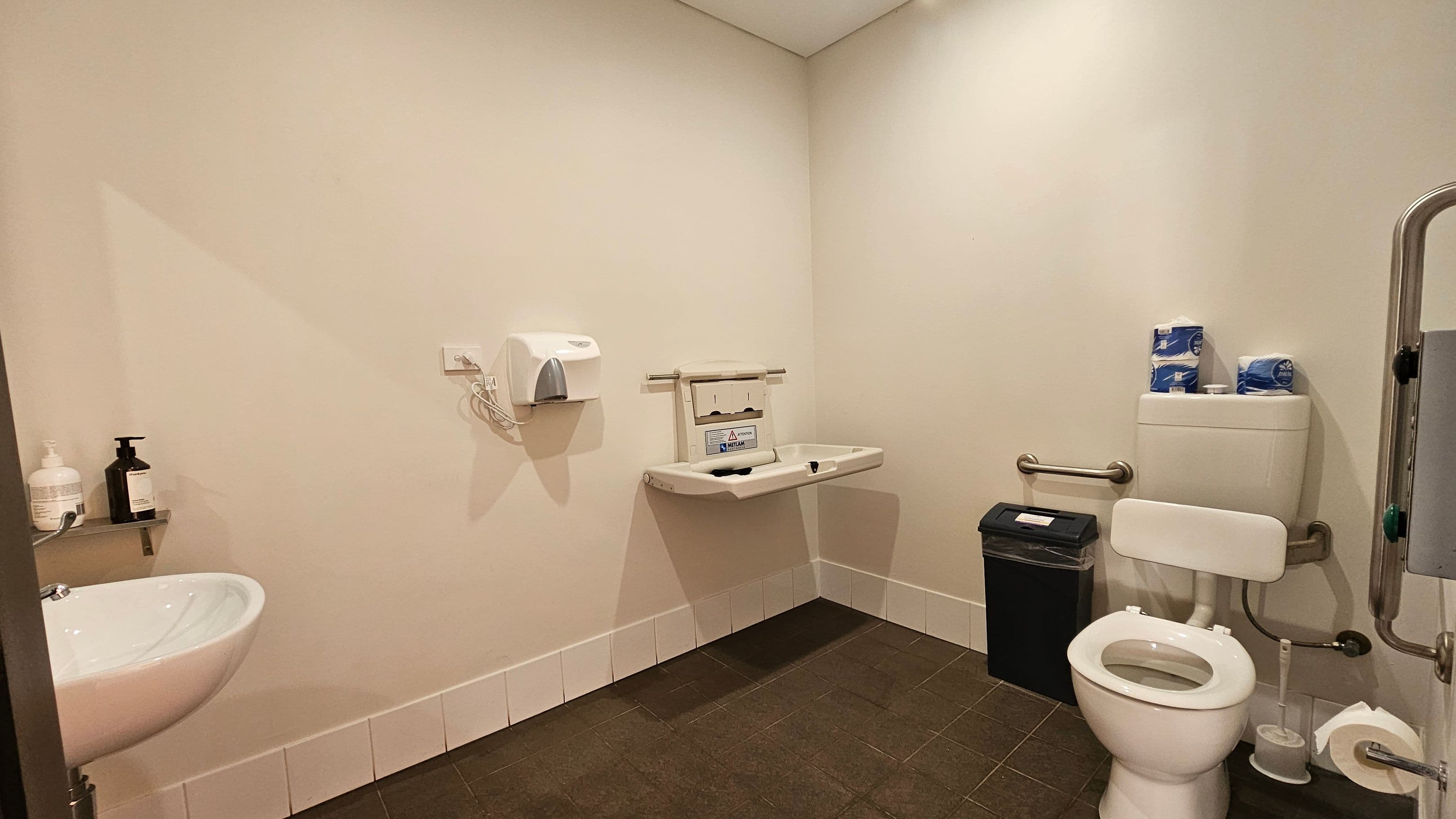 A public restroom with a white toilet on the right, equipped with a support rail and a black bin nearby. Above the toilet are rolls of toilet paper. In the centre, a baby changing station is mounted on the wall. On the left, a white sink with two soap dispensers sits below a hand dryer. The floor is dark tiled, and the walls are light-coloured.