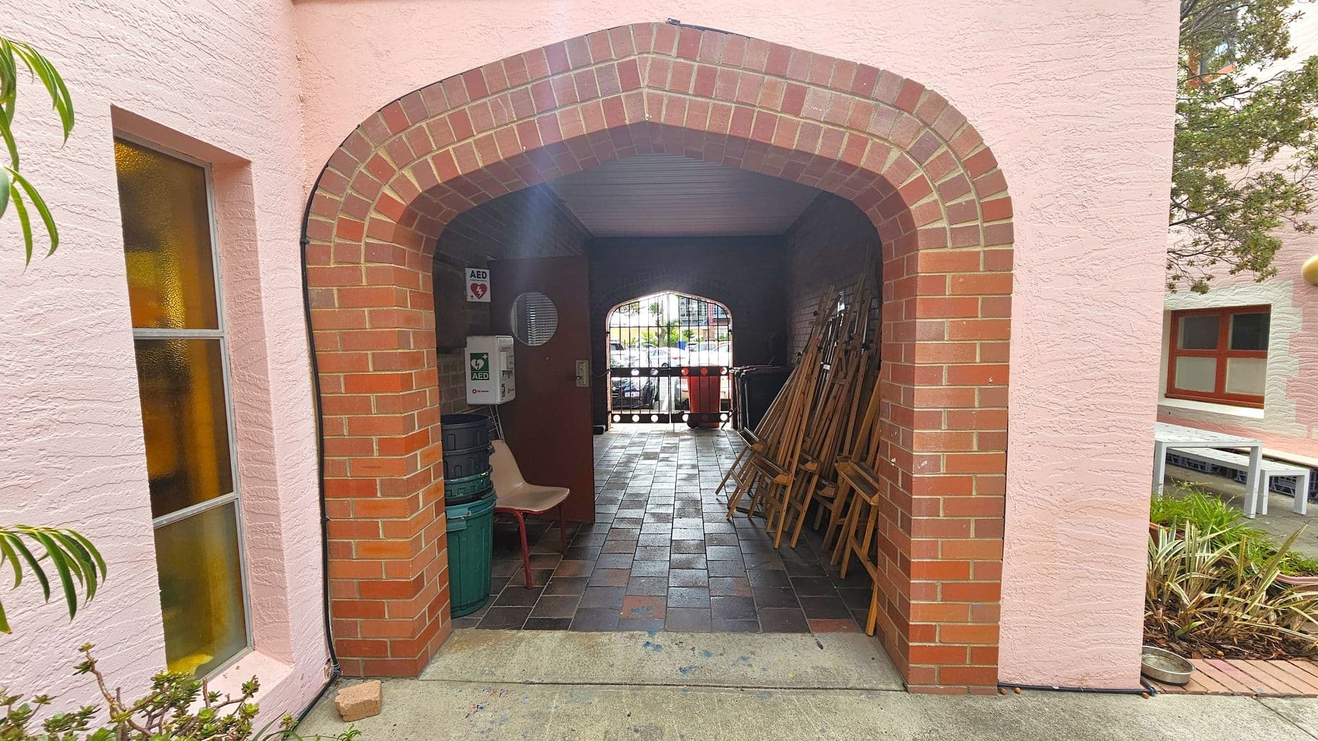 A brick archway leads into a covered passage with a tiled floor. On the left, there's a pink wall with a window featuring frosted glass. Inside the passage, a chair and some stacked bins are visible. On the right, several wooden easels are leaning against the wall. At the far end, a metal gate opens to an outdoor area with parked cars. The exterior walls are pink, and there are plants and a bench visible on the right side outside the archway.
