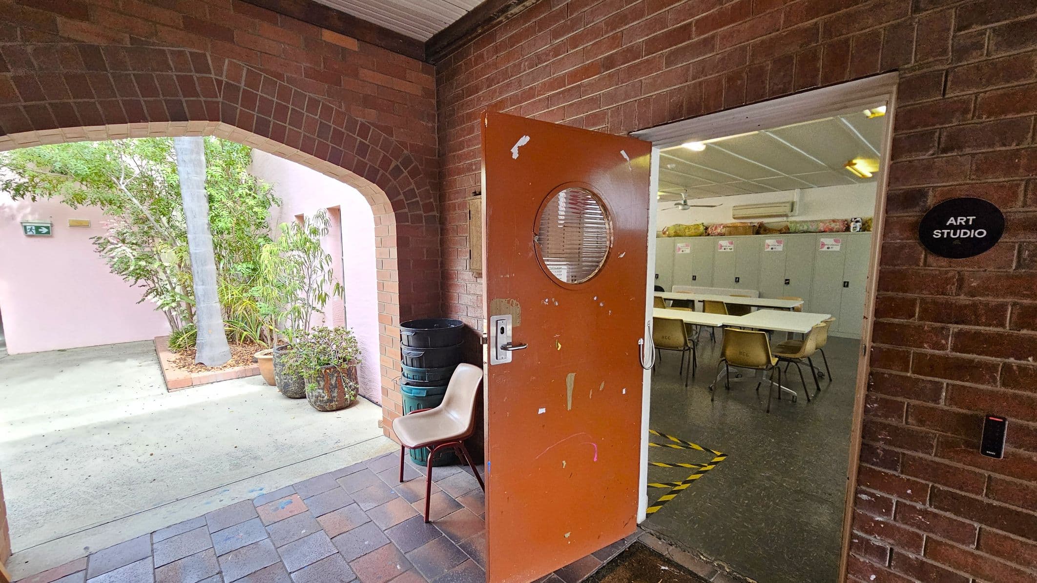 A brick archway on the left leads to an outdoor area with potted plants and a pink wall. In the centre, an open orange door with a round window leads into an art studio. Inside, there are tables, chairs, and lockers along the back wall. A sign on the wall reads "Art Studio". The floor outside is tiled, and the interior floor is dark with a yellow and black caution tape marking. A beige chair and stacked bins are near the door.