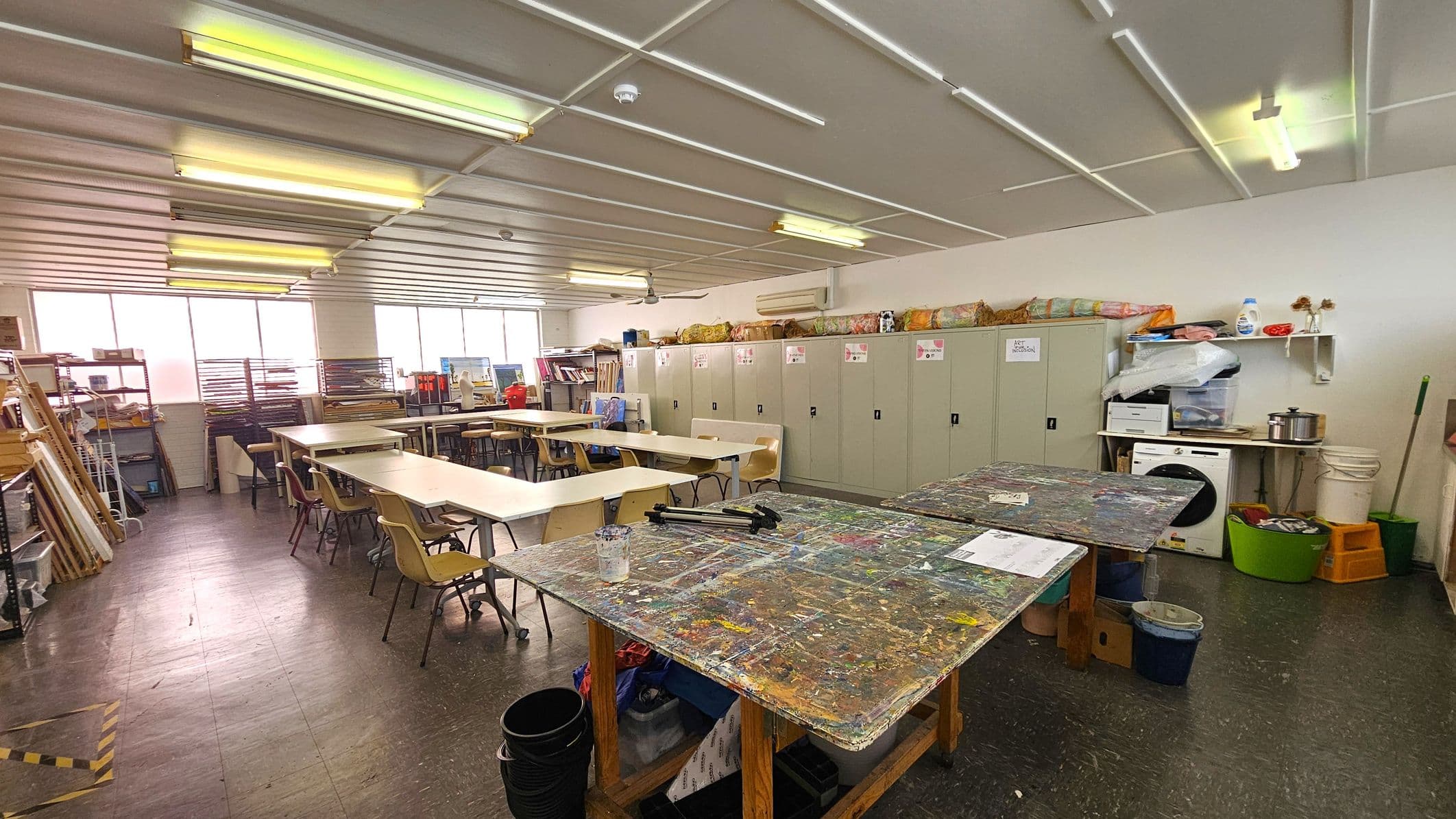 An art classroom with a cluttered, paint-splattered table in the foreground. To the right, there are storage cabinets with various art supplies on top. Shelves hold additional materials, including a printer and cleaning supplies. In the background, several tables and chairs are arranged for students. Large windows on the left let in natural light, and the ceiling has fluorescent lights. The floor is dark and speckled.