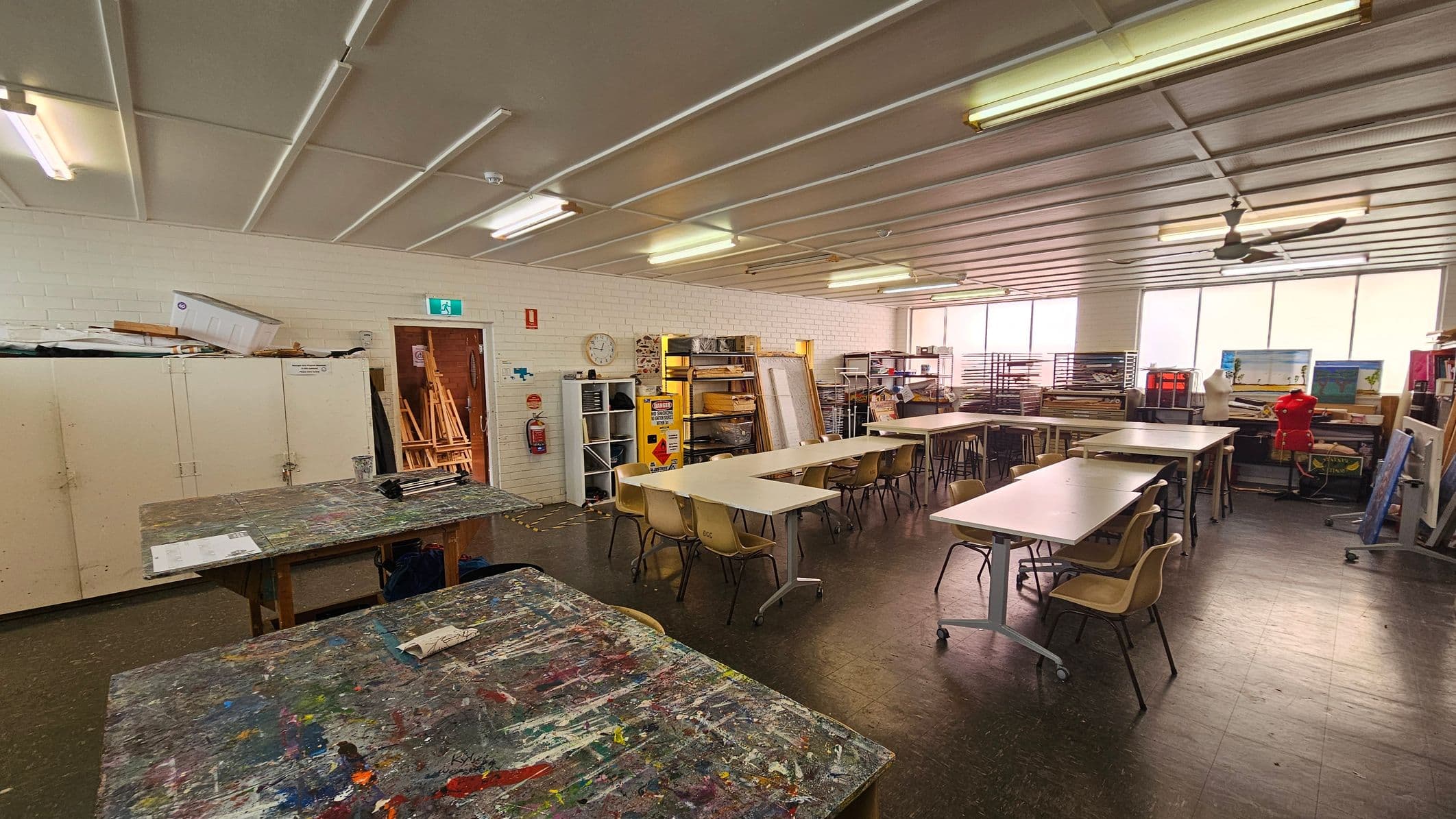 An art classroom with a cluttered, paint-splattered table in the foreground. To the left, there are white cabinets with items stacked on top. The middle features several tables and chairs arranged in rows. The right side has shelves with art supplies, a mannequin, and a red dress form. The back wall has large windows allowing natural light. The ceiling has fluorescent lights and a ceiling fan, and the floor is dark and tiled.