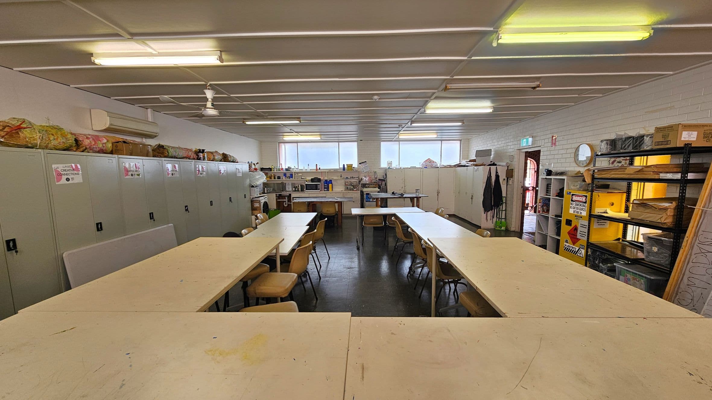 A room with a U-shaped arrangement of beige tables and brown chairs. On the left, there are grey lockers with items stored on top. The back wall has windows and shelves with various supplies, including a microwave. On the right, there are metal shelves with boxes and a "Danger No Smoking" sign. The ceiling has fluorescent lights and a fan, and the floor is dark.