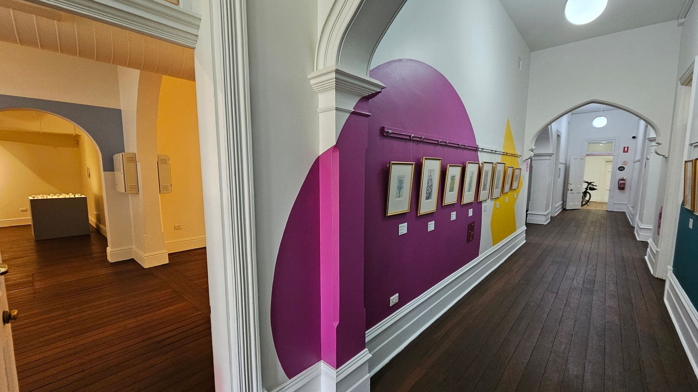 A hallway with dark wooden floors and white walls featuring colourful geometric patterns. On the right, framed artworks are displayed on a wall with pink and yellow sections. The ceiling is white with arched doorways. On the left, an adjacent room with light blue and yellow walls contains a grey pedestal. In the background, a door is open, revealing a bicycle and a fire extinguisher on the wall.