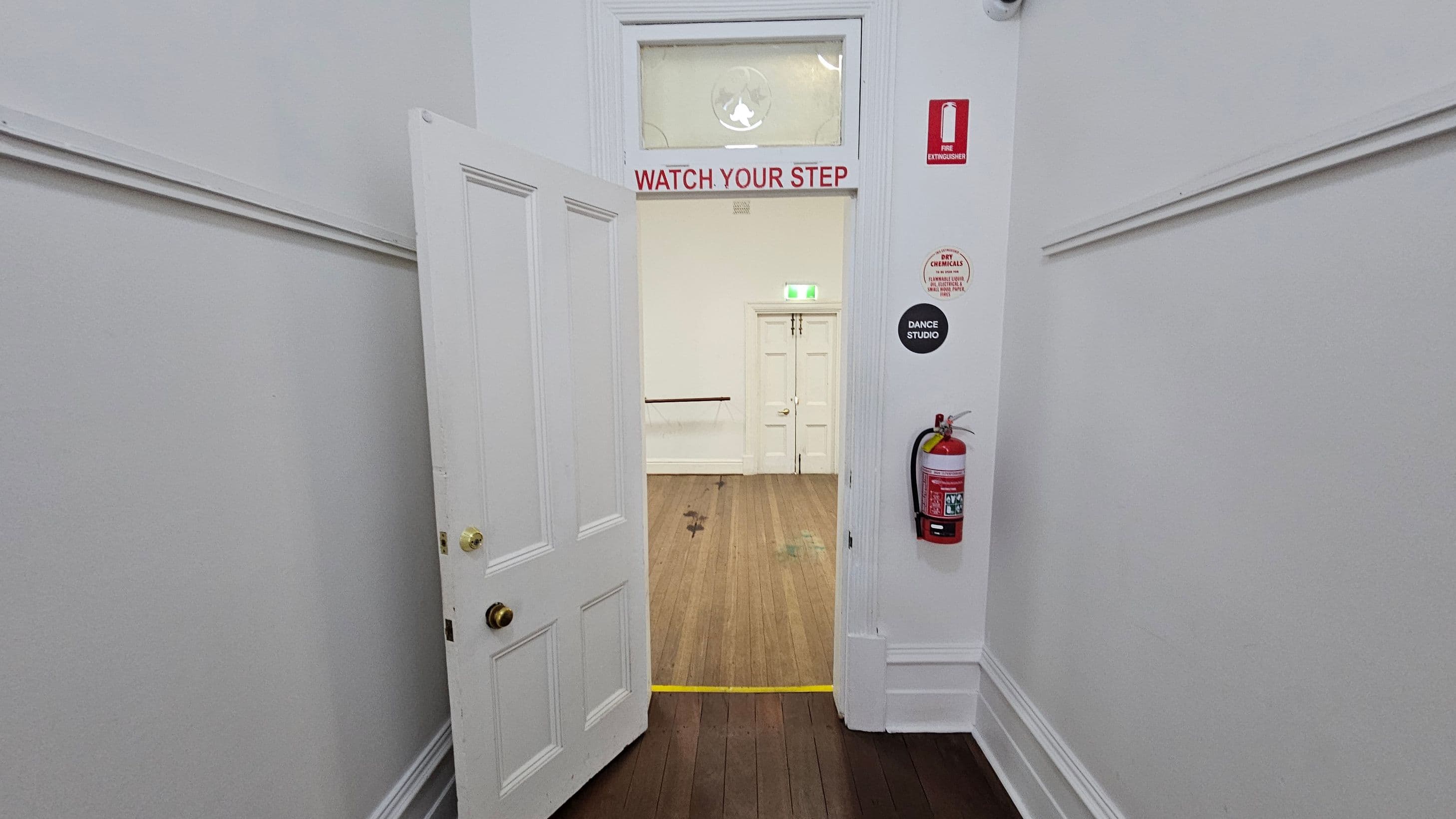 A white door is partially open, leading into a room with wooden flooring. Above the doorway, a sign reads "WATCH YOUR STEP" in red letters. On the right wall, there is a fire extinguisher and several signs, including one labelled "DANCE STUDIO". The room beyond has a white double door and a green exit sign above it. The walls are painted white, and there is a yellow line on the floor marking a step.