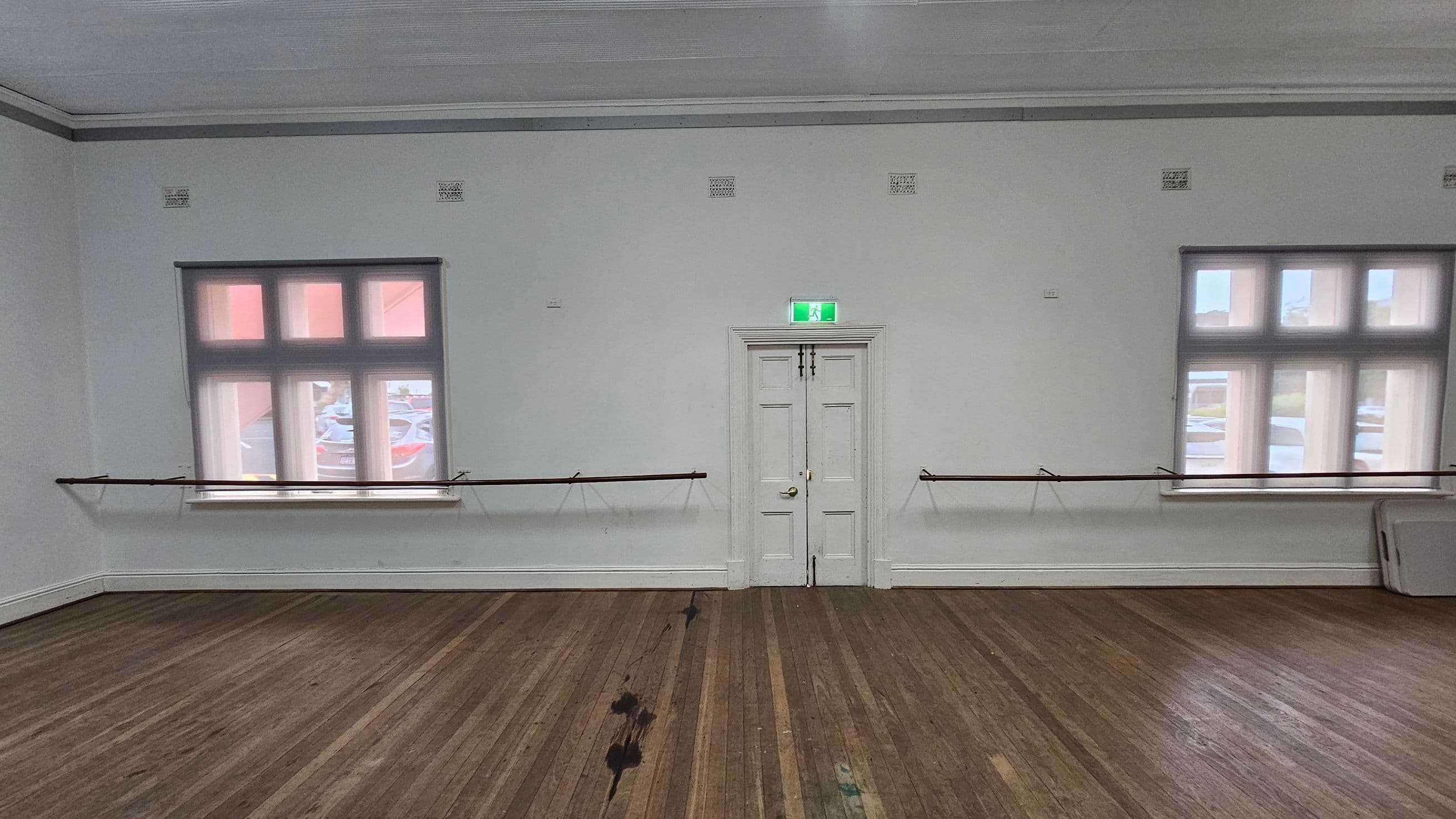 A room with a wooden floor and a white ceiling. In the centre, there is a white double door with an illuminated green exit sign above it. On either side of the door, there are two large windows with multiple panes, allowing light to enter. Along the wall, there are horizontal bars, possibly for ballet or exercise. A folded table leans against the wall on the right side. The floor has some dark marks and a purple stain.