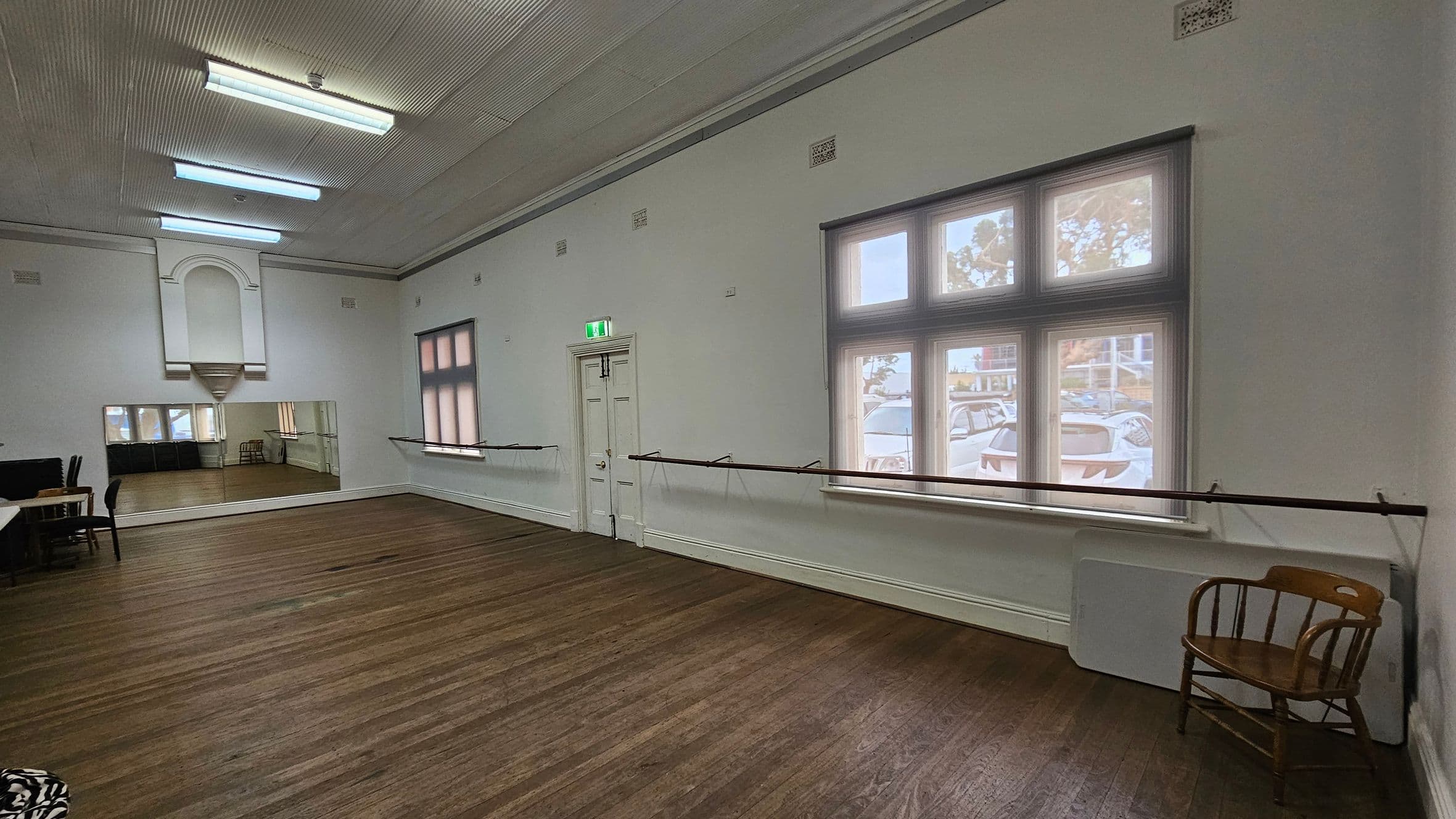 A spacious room with wooden flooring and a high ceiling featuring fluorescent lights. On the right, large windows with a view outside and a wooden chair beneath them. A ballet barre runs along the wall. On the left, a large mirror reflects the room, and there are tables and chairs. A door with an exit sign is centred on the back wall. The walls are painted white, and the room has a clean, open feel.