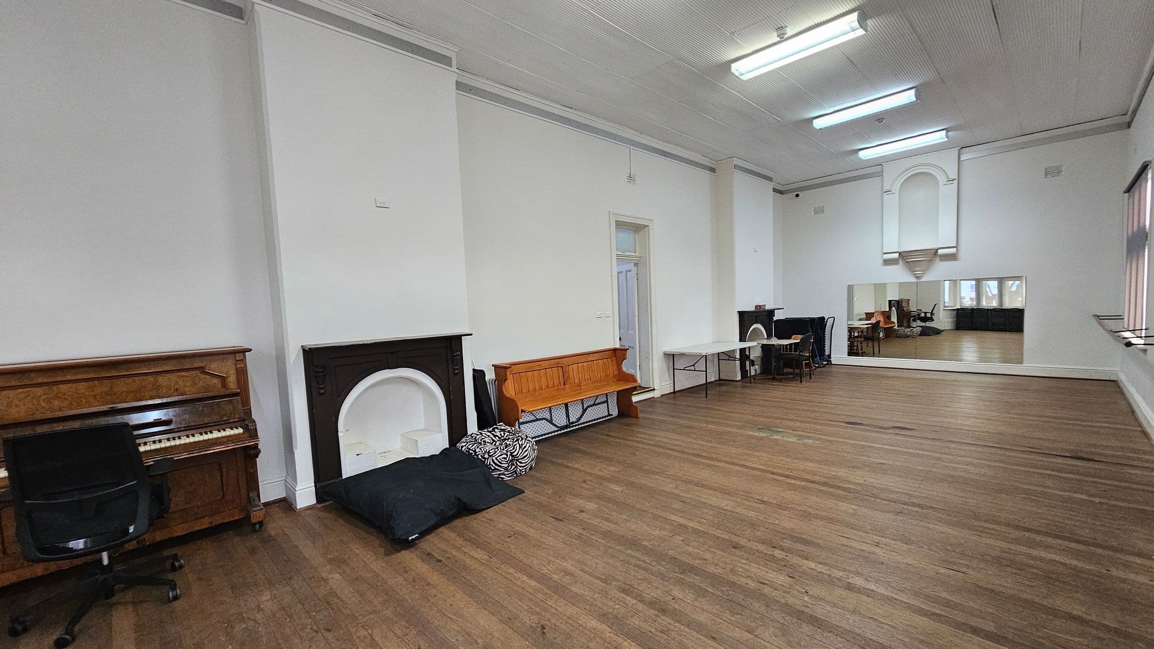 A spacious room with wooden flooring and a high ceiling with fluorescent lights. On the left, there's an upright piano with a black office chair in front of it, next to a black fireplace. A wooden bench is placed against the wall with cushions on the floor nearby. In the middle, a white door is visible. On the right, a large mirror reflects the room, and a table with chairs is set up near another fireplace. Large windows with blinds line the right wall.