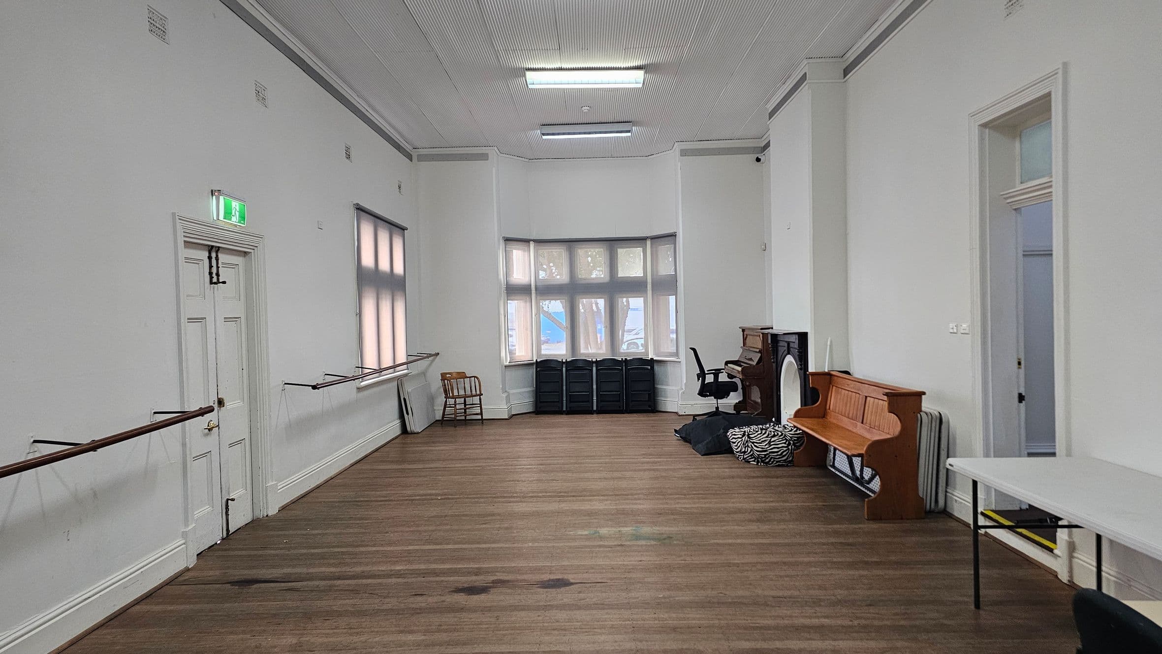 A spacious room with wooden flooring and a high, white panelled ceiling. On the left, a white door with an exit sign above it is flanked by ballet barres. Large windows with blinds are on the left wall. At the far end, a bay window with multiple panes is partially covered by black storage units. On the right, a wooden bench, a piano, and a chair are near a doorway. A white table is positioned on the right side.