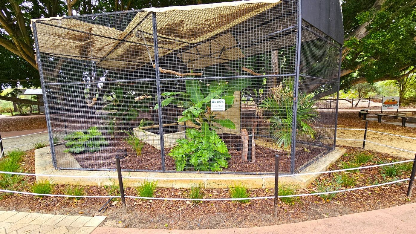 A large, hexagonal wire enclosure with a mesh roof, housing various plants and branches inside. A sign on the front reads "WE BITE! Do not put fingers through the wire." The enclosure is surrounded by a low stone border with small plants and a rope barrier. In the background, there are trees and a sign that reads "WILDLIFE PARK." The ground is covered with mulch and a paved path runs alongside the enclosure.