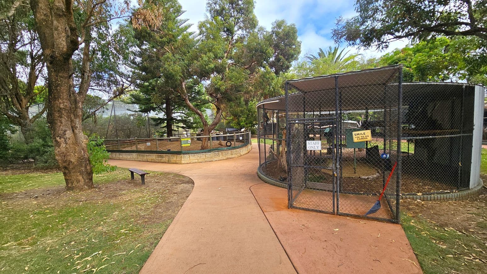 A pathway curves through a park area. On the left, there's a large tree with a bench nearby. In the centre, a fenced enclosure with a sign reading "Staff Only" and another saying "Smile, you're on camera" is visible. The enclosure has a metal mesh and a broom leaning against it. In the background, more trees and greenery are present, with a wooden fence enclosing another area. The ground is a mix of grass and a paved path.