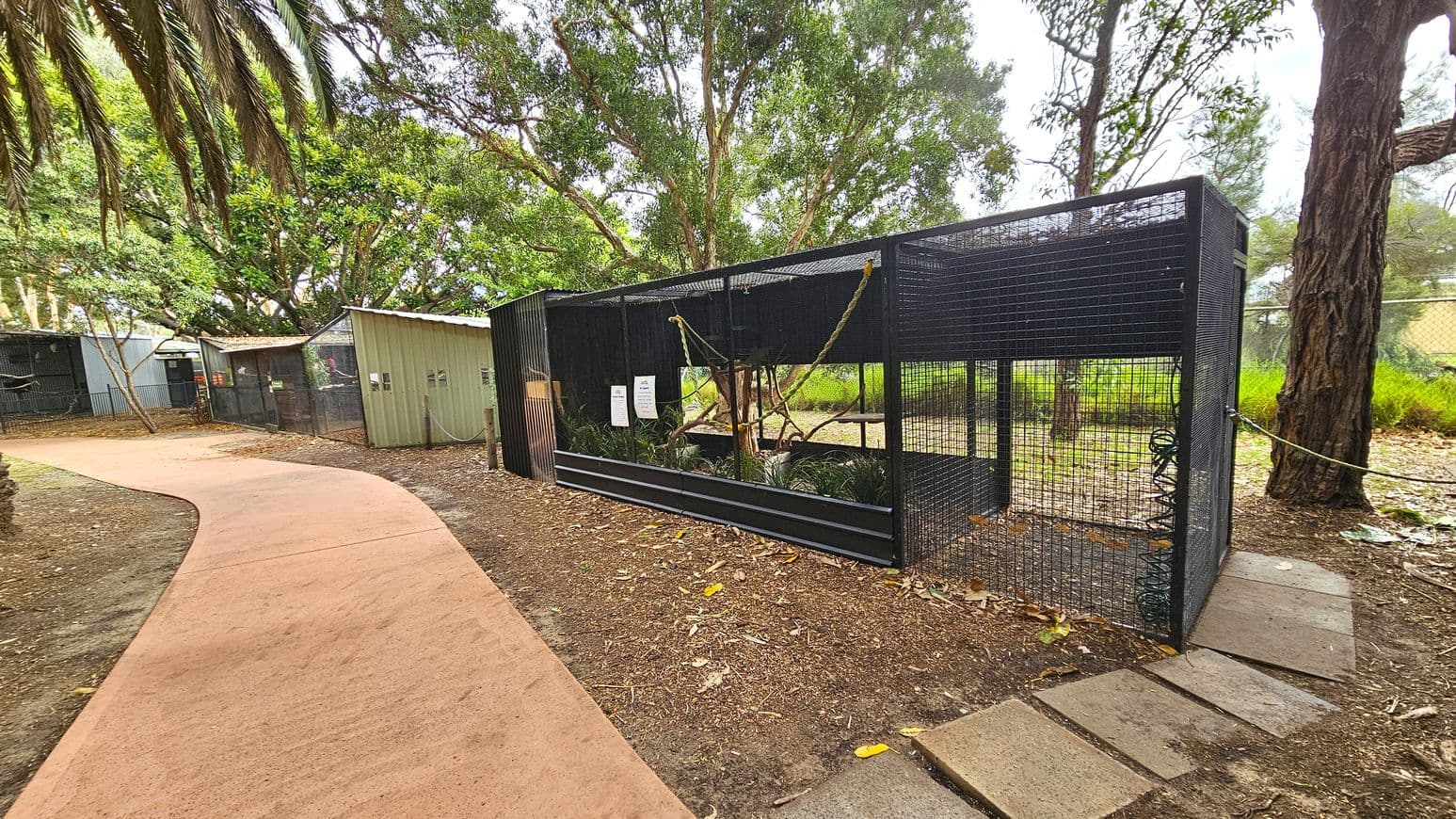 A pathway curves through a wooded area, bordered by a series of enclosures. On the right, a large black mesh cage contains plants and branches, with signs attached to the front. To the left, smaller green structures with corrugated roofs are visible. The ground is covered with mulch and fallen leaves, and tall trees with dense foliage provide shade. A rope barrier runs alongside the path, and the background features a grassy area and more trees.