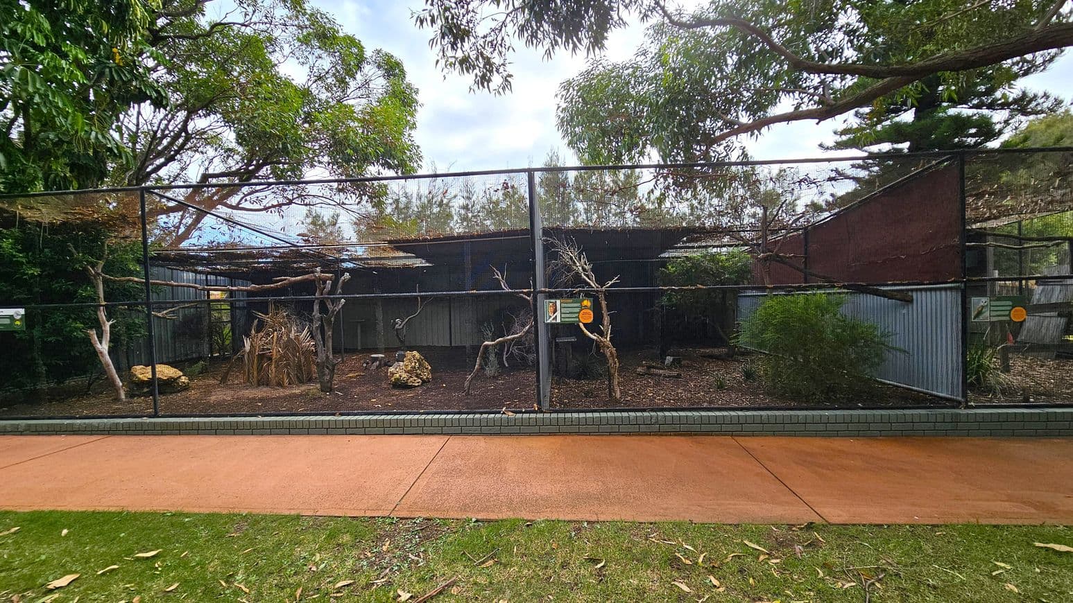 A large outdoor aviary with a wire mesh enclosure. Inside, there are several dry branches and rocks scattered on the ground, with some sparse vegetation. The background features corrugated metal walls and a partial roof. Trees with green foliage surround the aviary, and a paved path runs along the front, bordered by grass. Signs are attached to the mesh, and the area is shaded by overhanging tree branches.