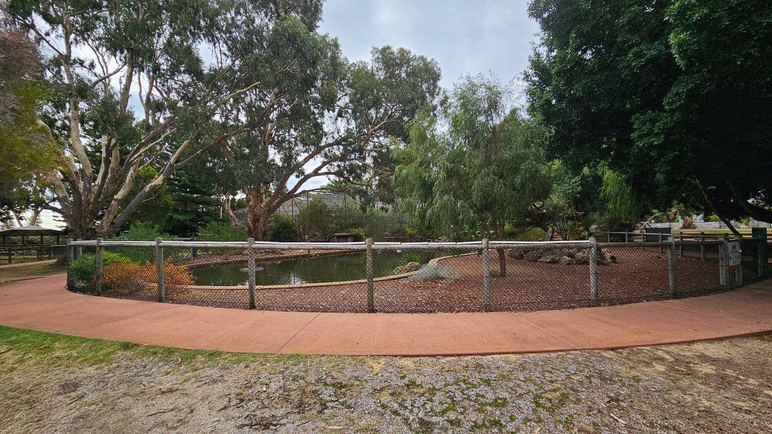 A fenced pond is in the centre, surrounded by a red paved path. Tall trees with dense foliage are in the background, providing shade. The ground in the foreground is a mix of grass and dirt. On the left, there is a small covered area with a bench. The right side has more trees and a sign near the fence.